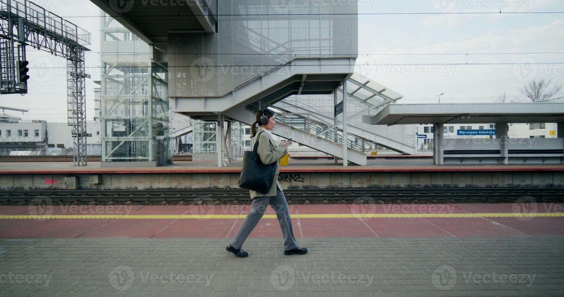 A woman in formal clothes and with headphones on her head uses a mobile phone walking along the railway platform - headphones photo