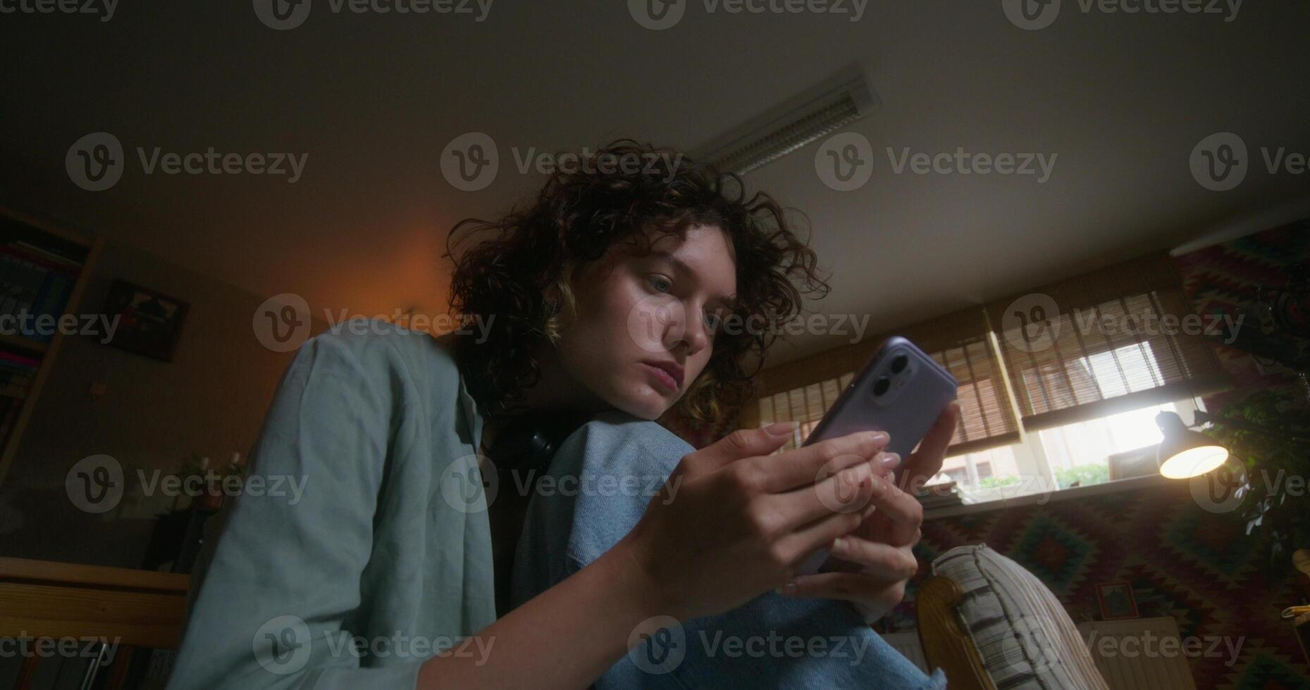 A young woman uses a mobile phone while sitting on a bed at home with a depressed look - mobile photo