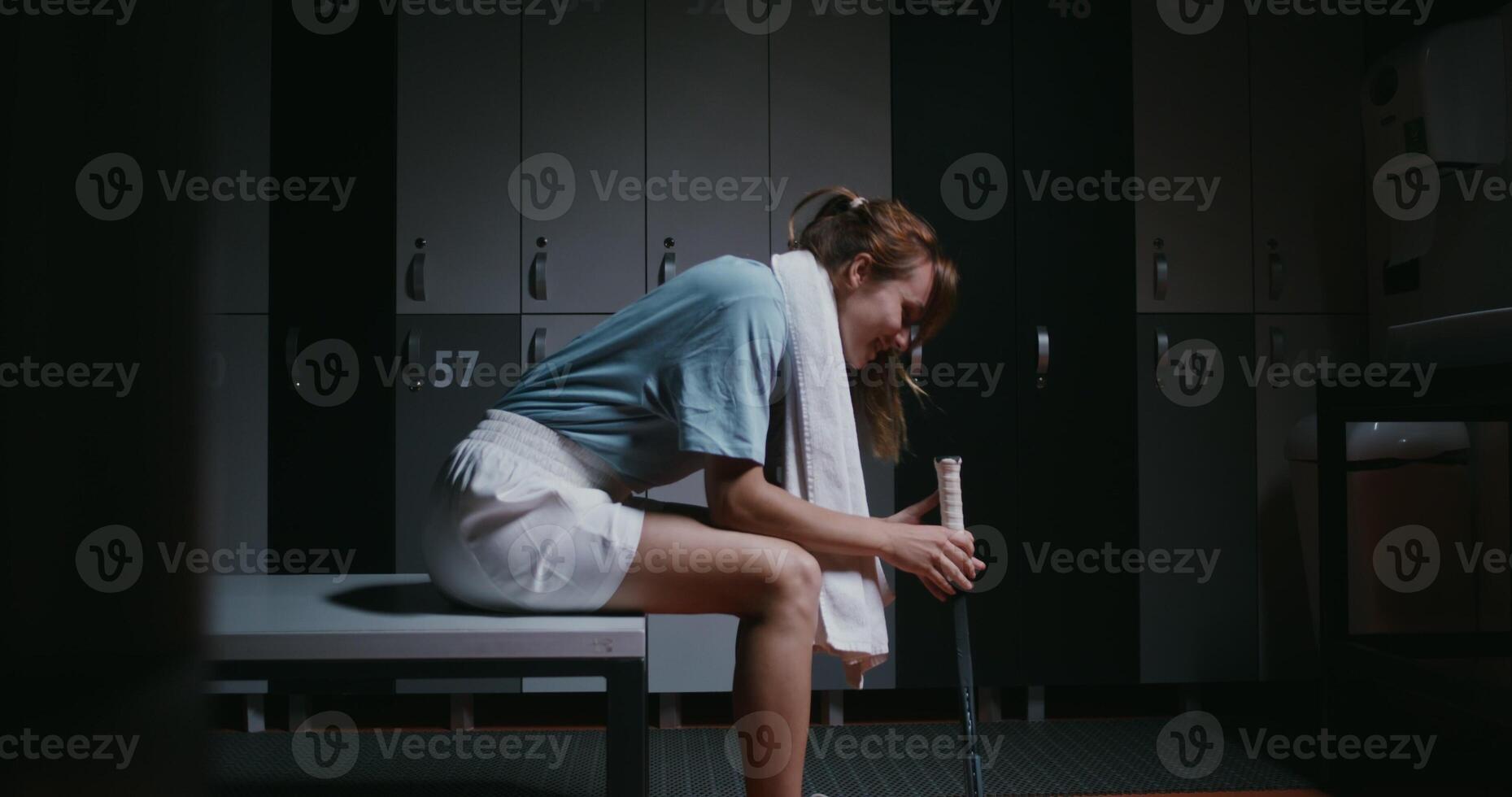 Young beautiful woman tennis player is resting in the locker room after the match, sitting on a bench with a towel around her neck and twirling a tennis racket in her hands. photo
