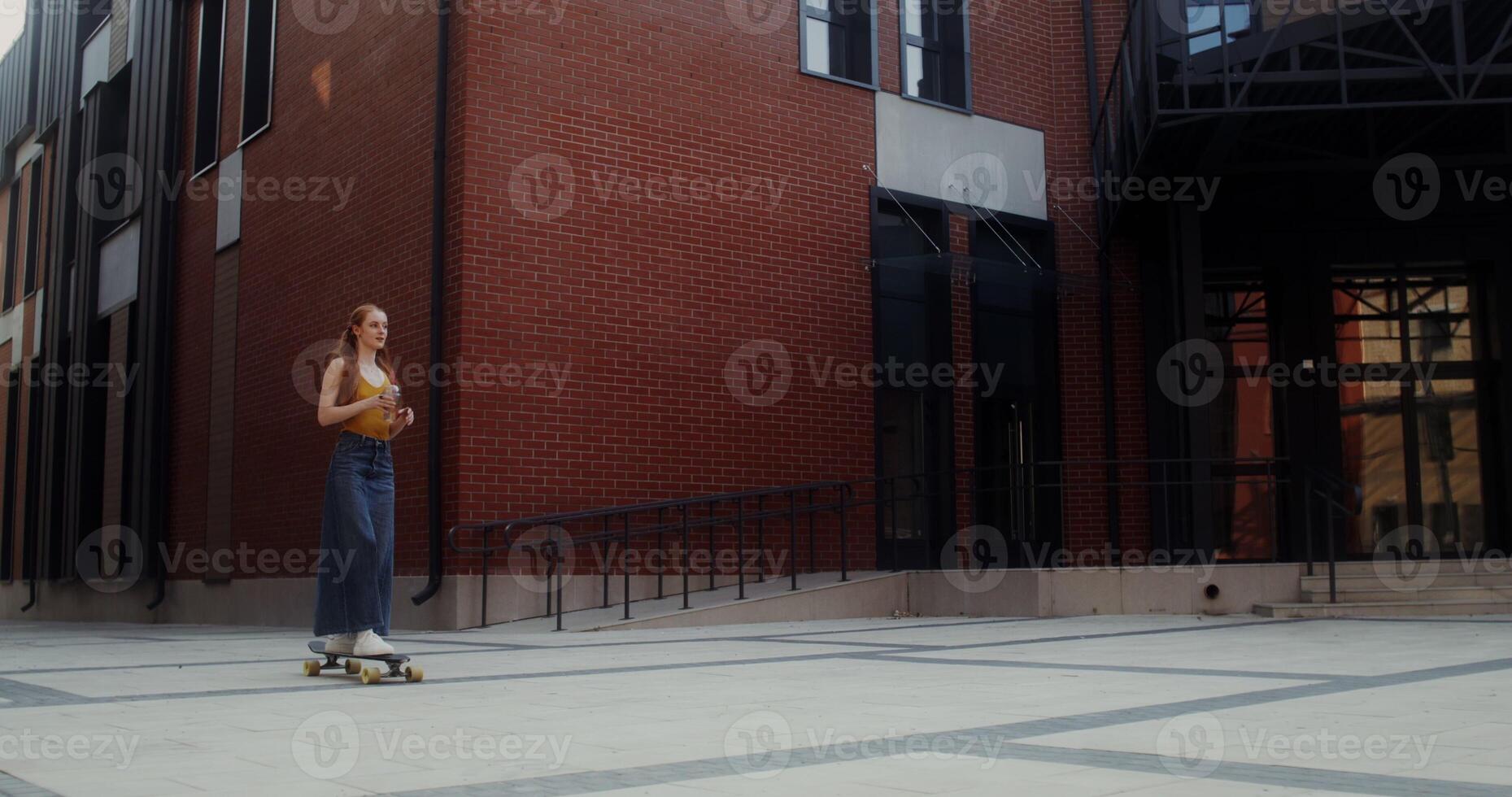 A young red-haired woman drinks lemonade while riding a longboard on a city street on a summer day - longboard photo