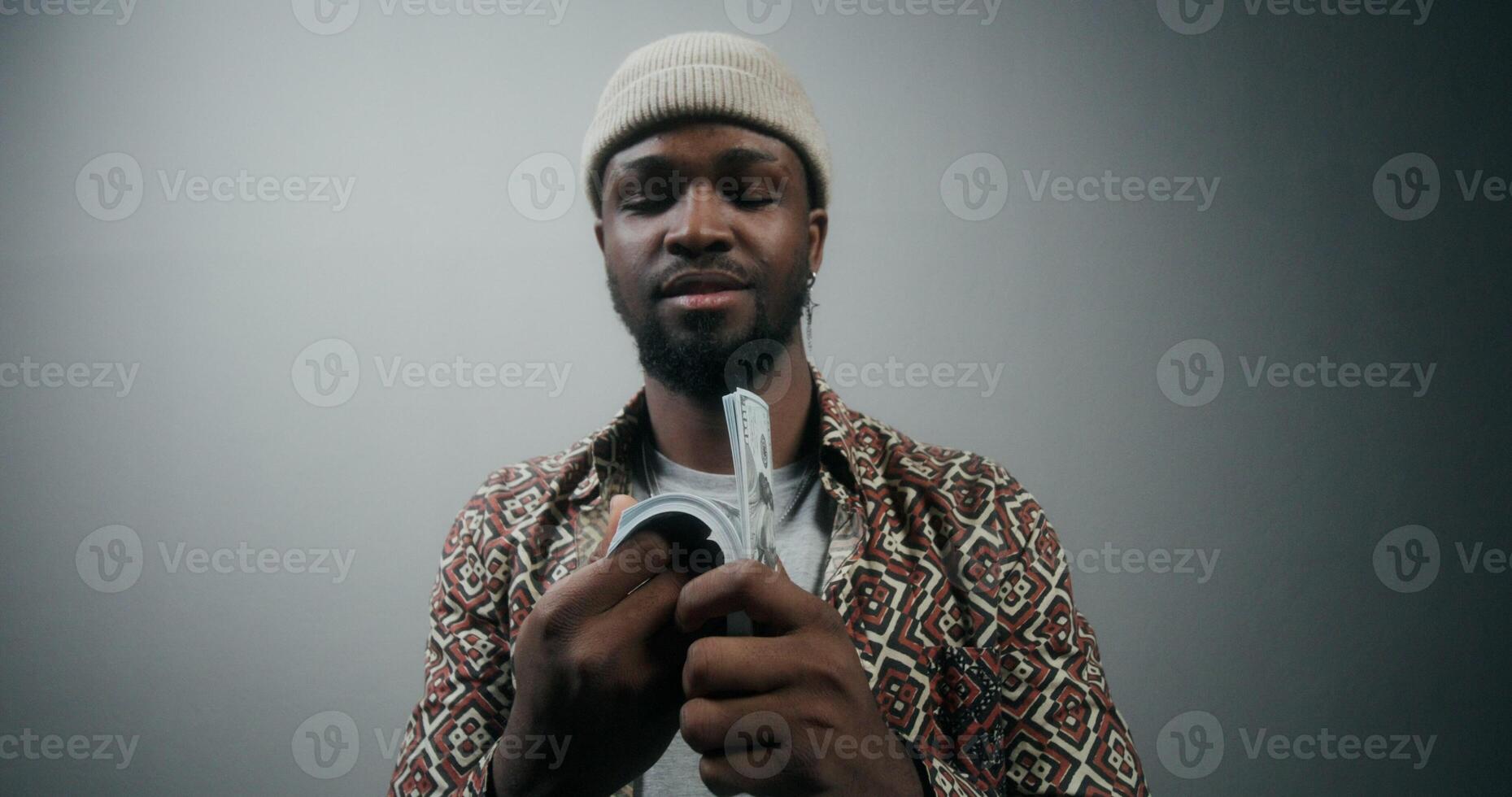A stylish bearded man in a hat tosses a stack of money and smiles at the camera standing on a gray background in the studio - tosses photo