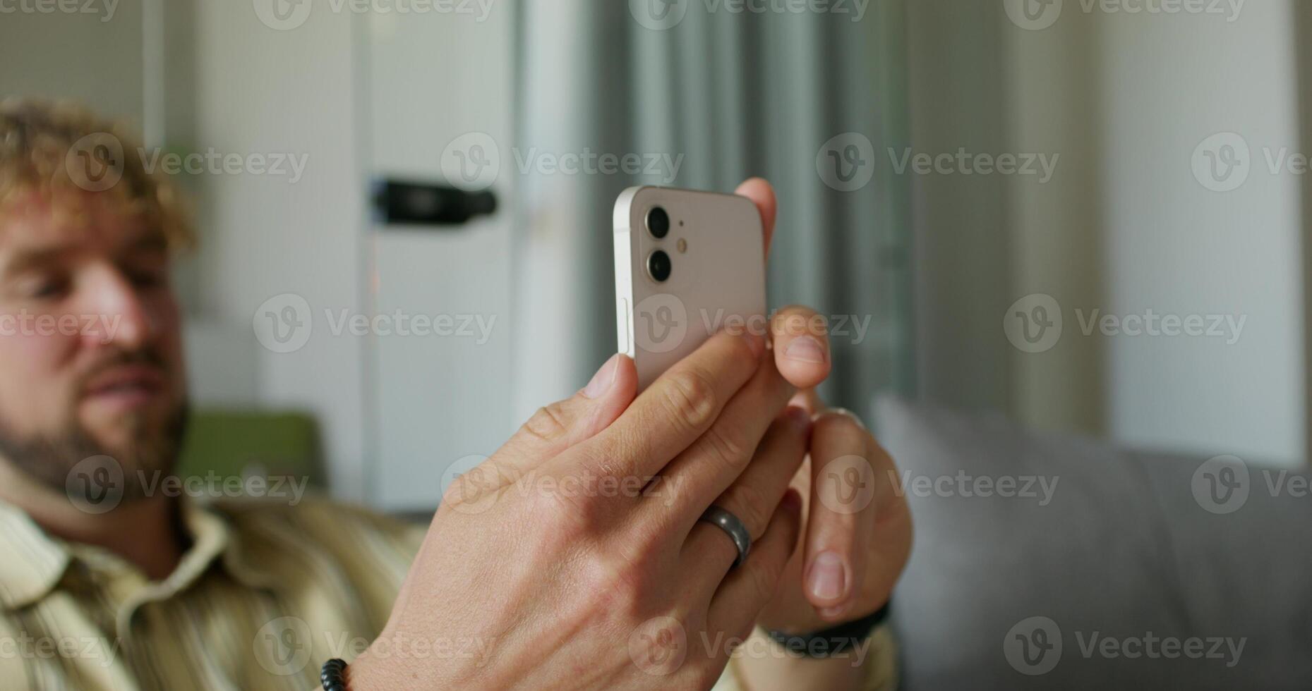 A bearded man in casual clothes is talking on a chat on a mobile phone sitting on the couch at home. Daylight photo