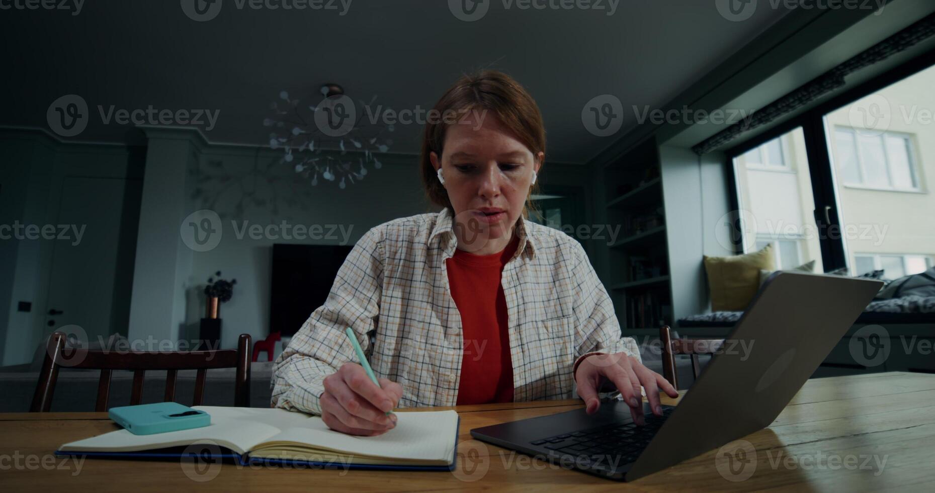 A young woman in casual clothes makes notes in a notebook sitting in front of an open laptop in a stylish home interior, with a serious look photo