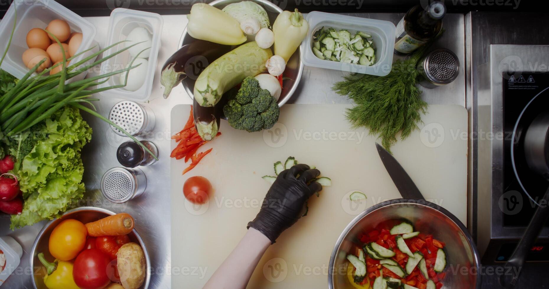 A woman in disposable black gloves adds a sliced cucumber to a salad bowl, standing near a table filled with vegetables, close photo