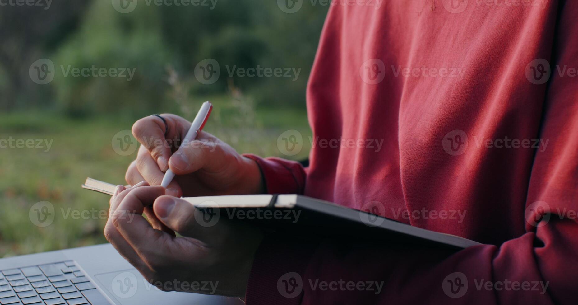 A young man of European appearance makes notes in a notebook, using a laptop, sitting on the ground under a tree, close-up photo