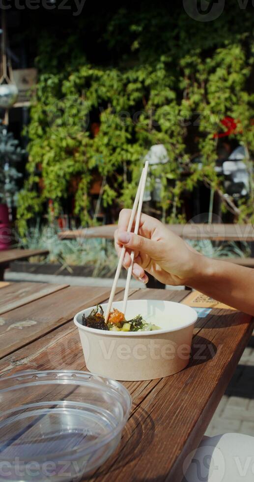 A young woman in casual clothes eats Asian food out of a box using chopsticks while sitting at a table on the cafe terrace - chopsticks photo