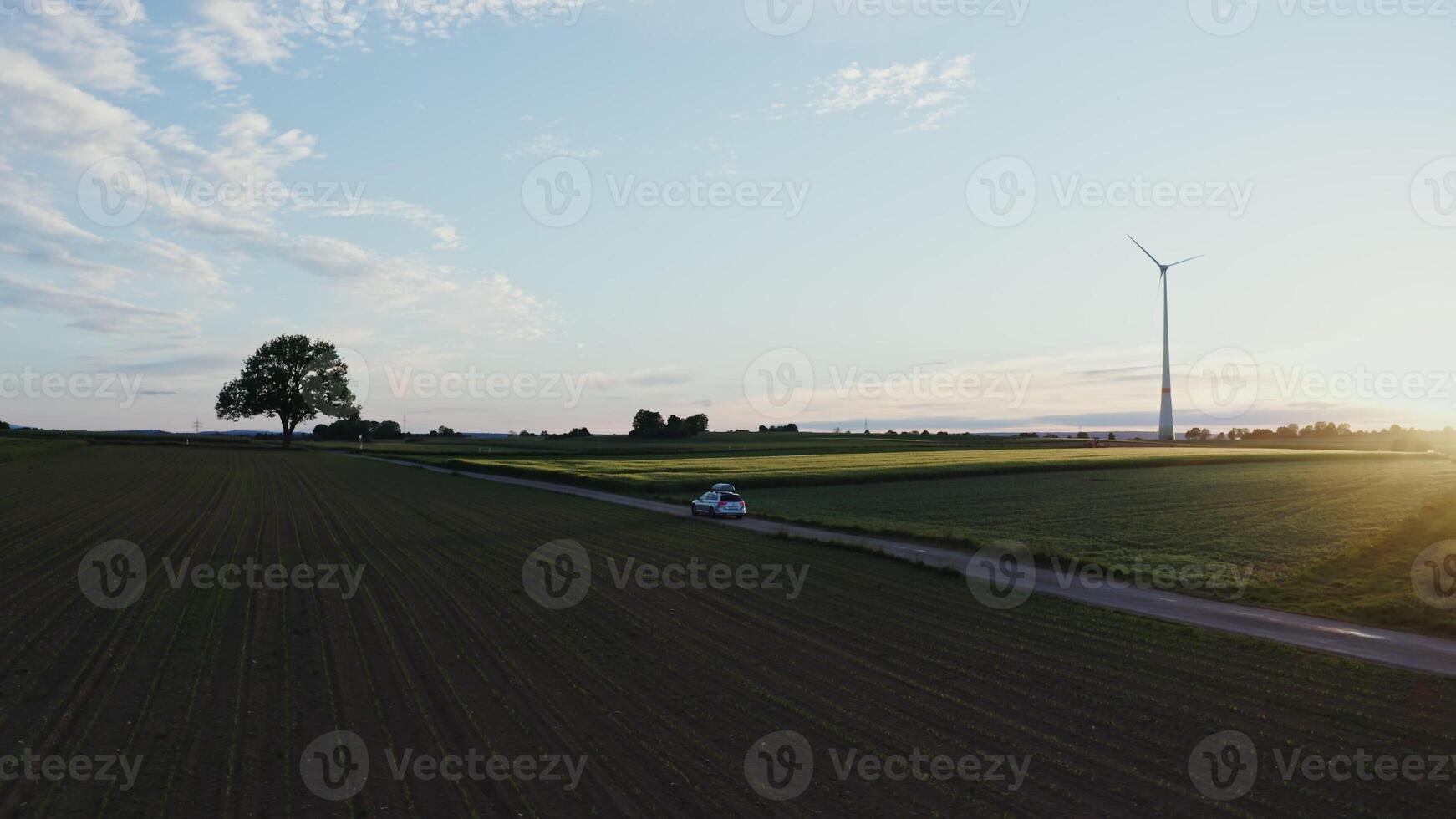 A car with a roof rack drives along a road through picturesque green meadows and arable fields on a beautiful sunny morning. Wind generator in the rays of the rising sun - meadows photo