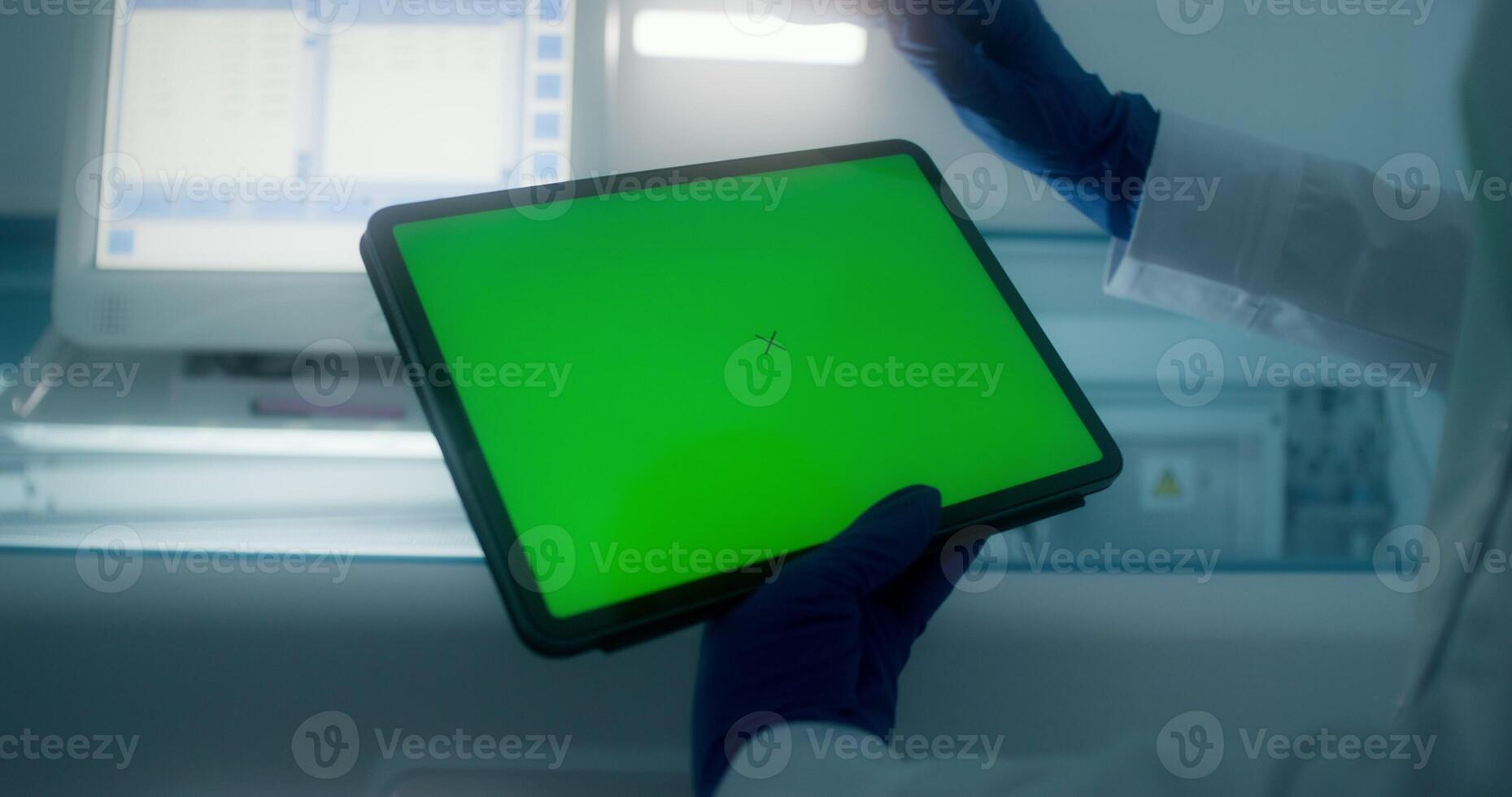 A woman in a lab coat and disposable gloves holds a tablet with a green screen standing in a medical laboratory. Close-up of her hands, copy space - screen photo