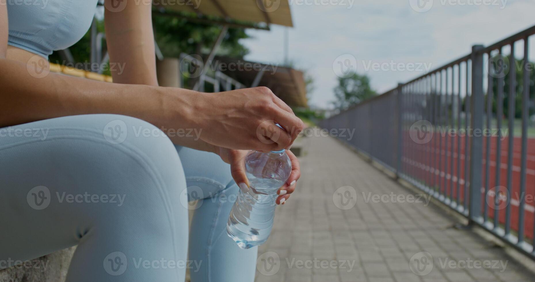 A woman in sports clothes and with earbuds in her ears is resting sitting on a stadium seat. She drinks water from a bottle. Summer day - earbuds photo