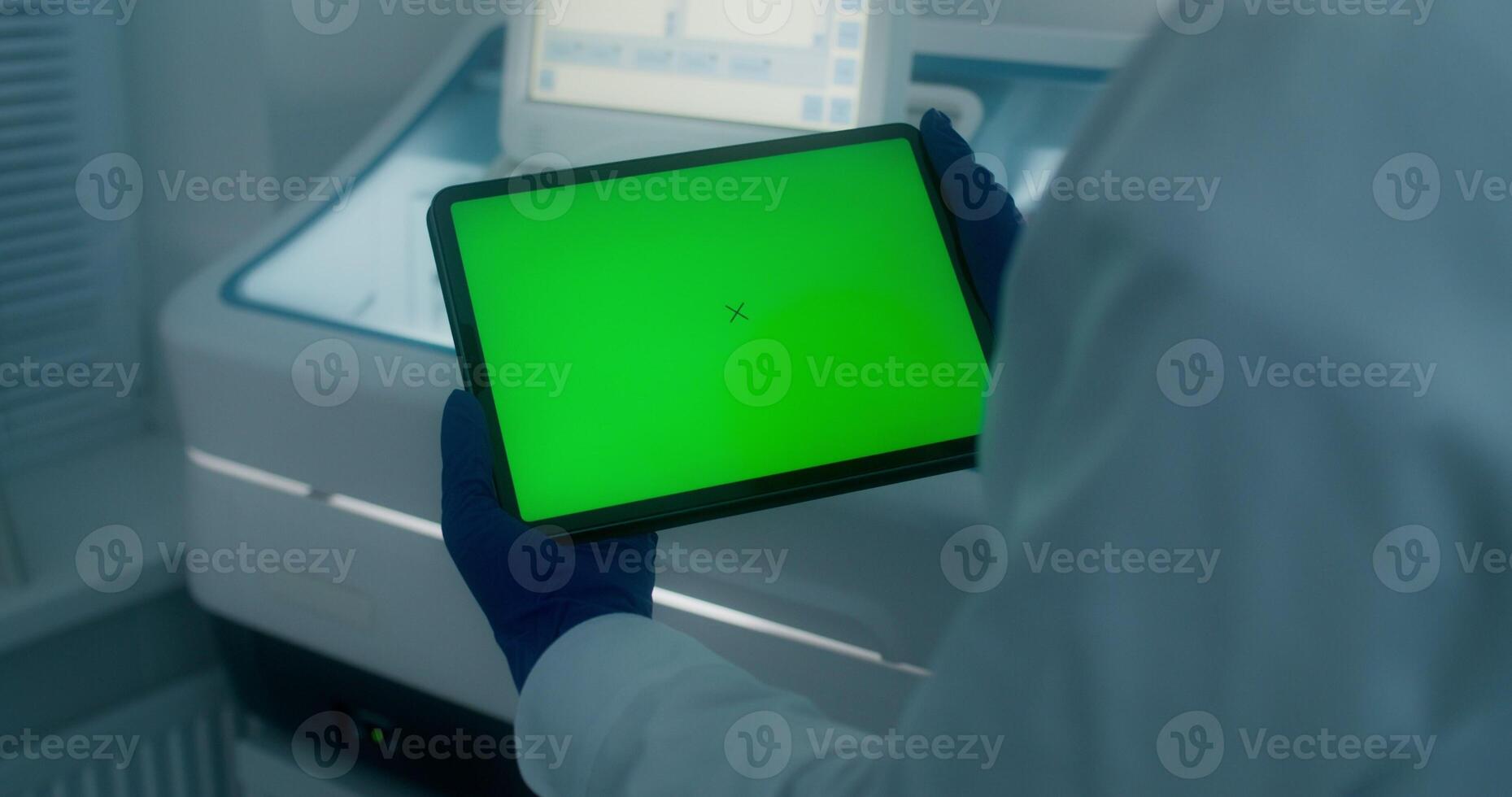 A woman in a lab coat and disposable gloves holds a tablet with a green screen standing in a medical laboratory. Close-up of her hands, copy space - tablet photo