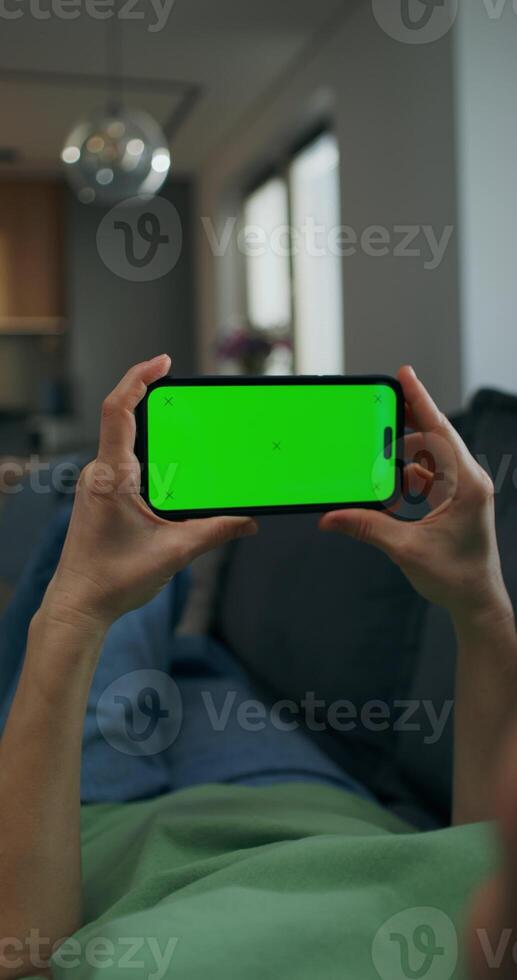 A woman holds a mobile phone with green screen in her hand. She is lying on the couch at home, close-up of her hands, unrecognizable person - person photo