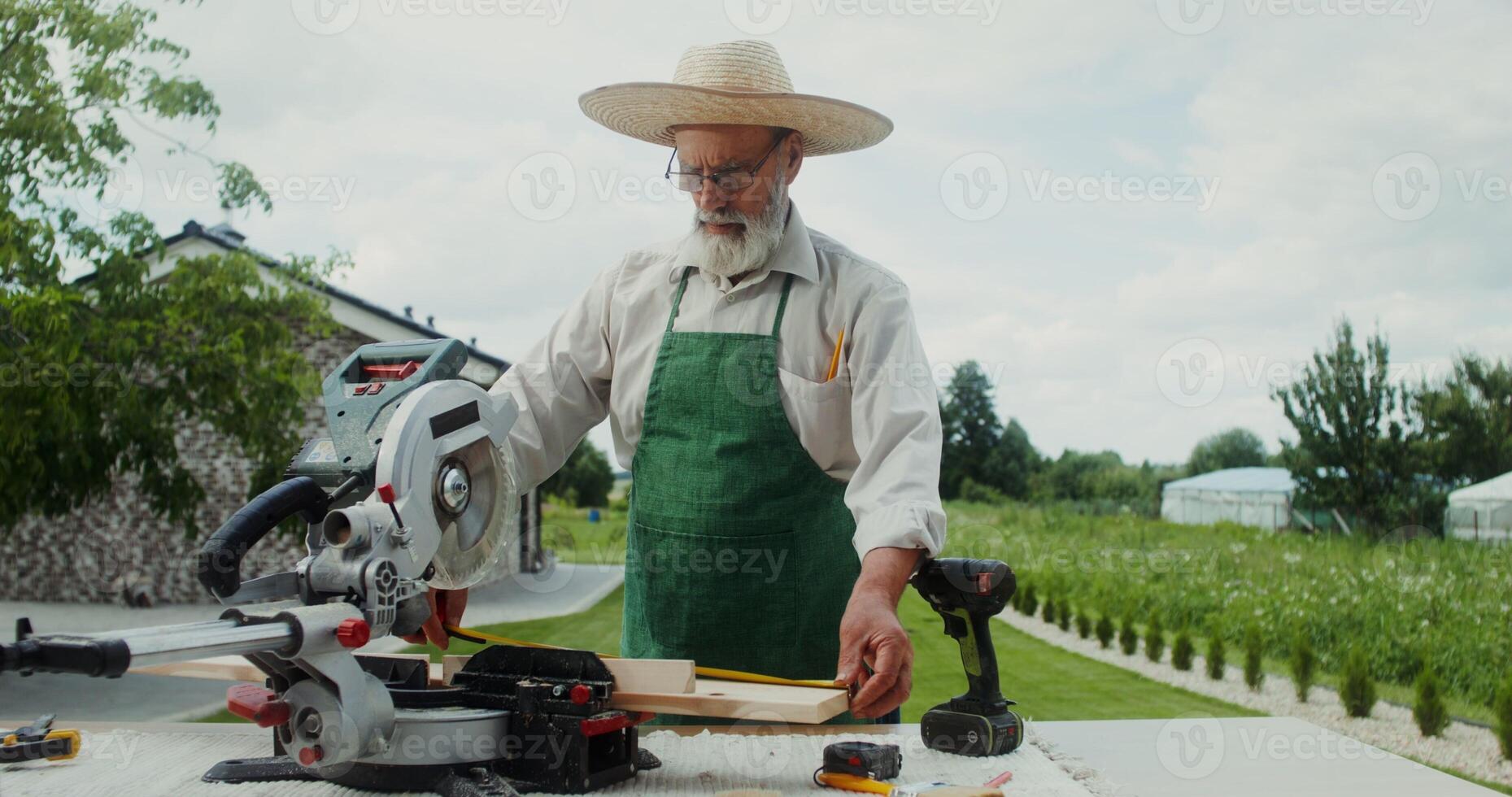 An elderly carpenter man in an apron and hat uses a tape measure standing near a miter saw in the backyard of his house - carpenter photo