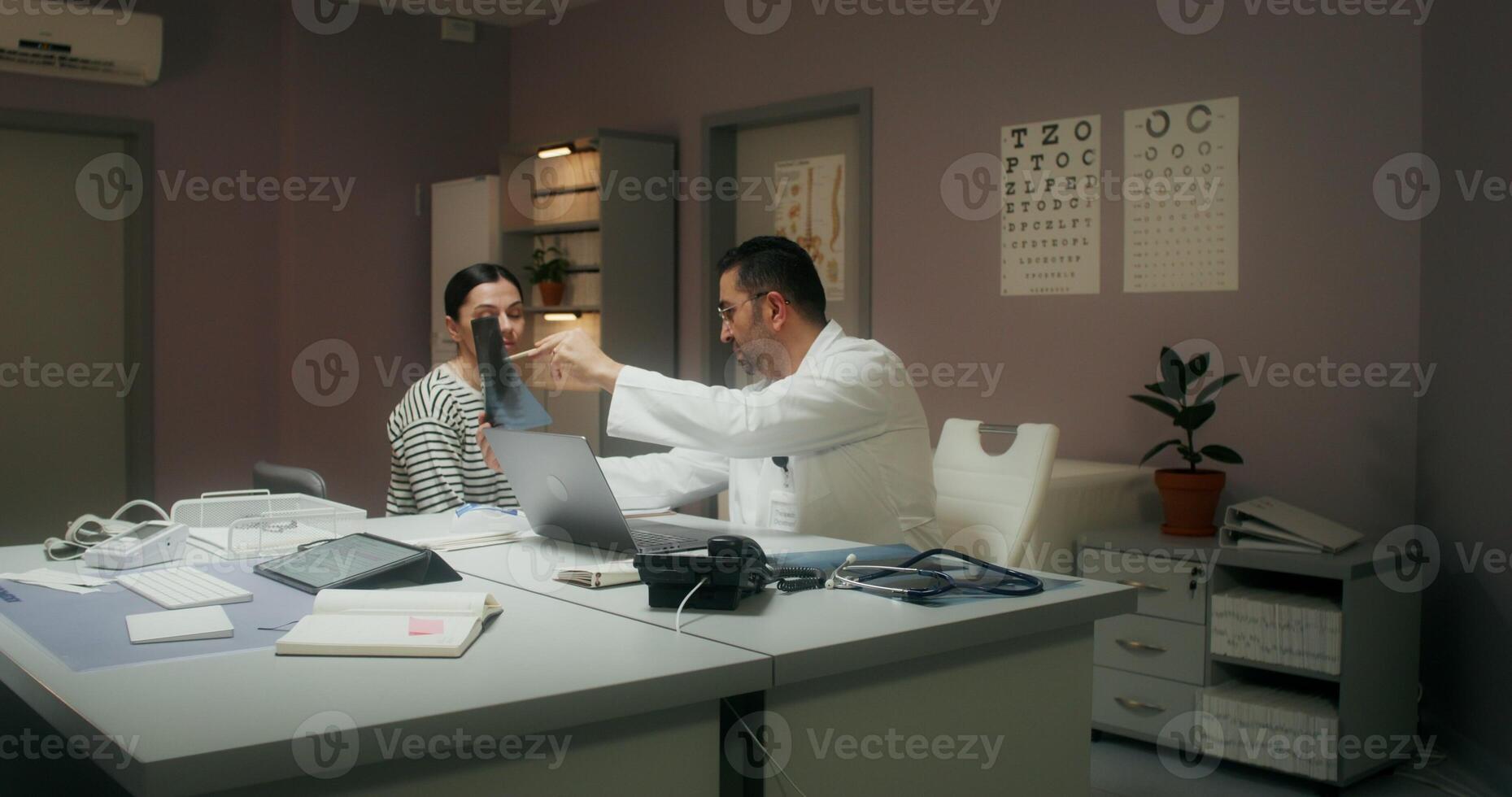 A doctor in a white coat shows a woman patient an X-ray, and then writes down appointments in a notebook, sitting at his desk in the medical office at the clinic - writes photo