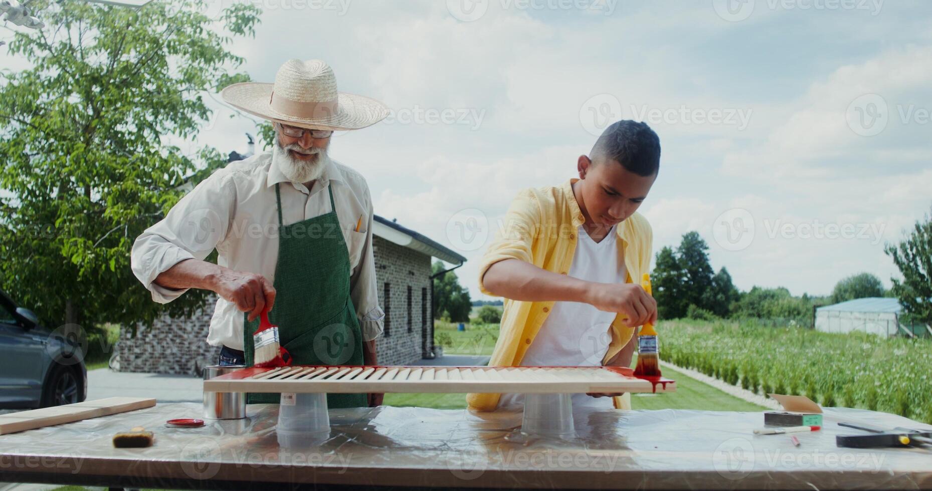 An elderly man in an apron and hat teaches a boy how to paint a door properly while standing in the backyard photo