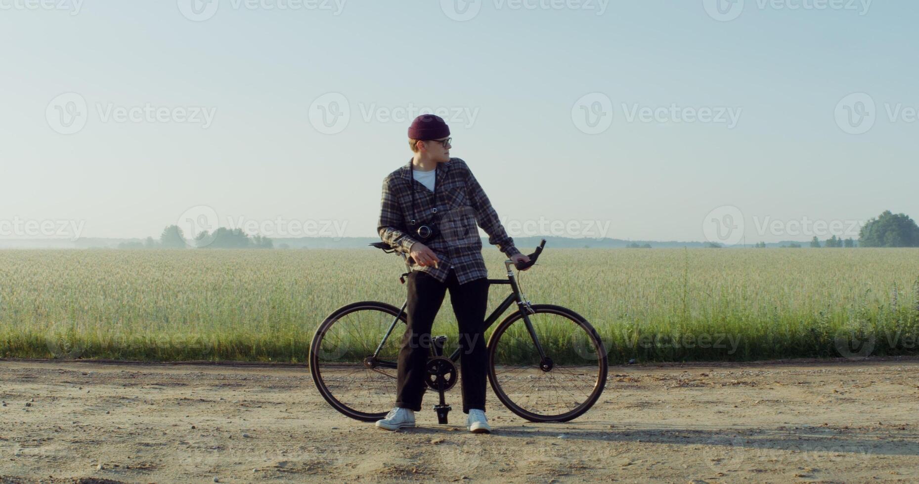 A young man with a camera around his neck stopped near a wheat field, resting after cycling and admiring the beauty of nature in the rays of the rising sun. 4k , red komodo photo