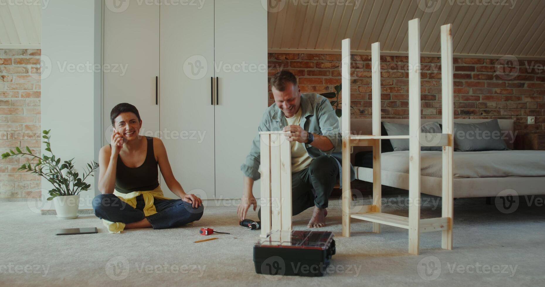 A woman is talking on a mobile phone sitting on the floor next to a man assembling a rack with a screwdriver, they are talking photo
