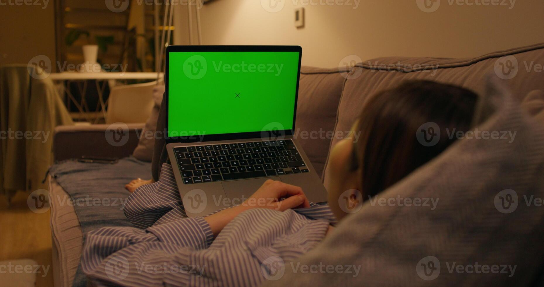 A woman uses a laptop with green screen lying on the couch in an apartment with dim lights photo