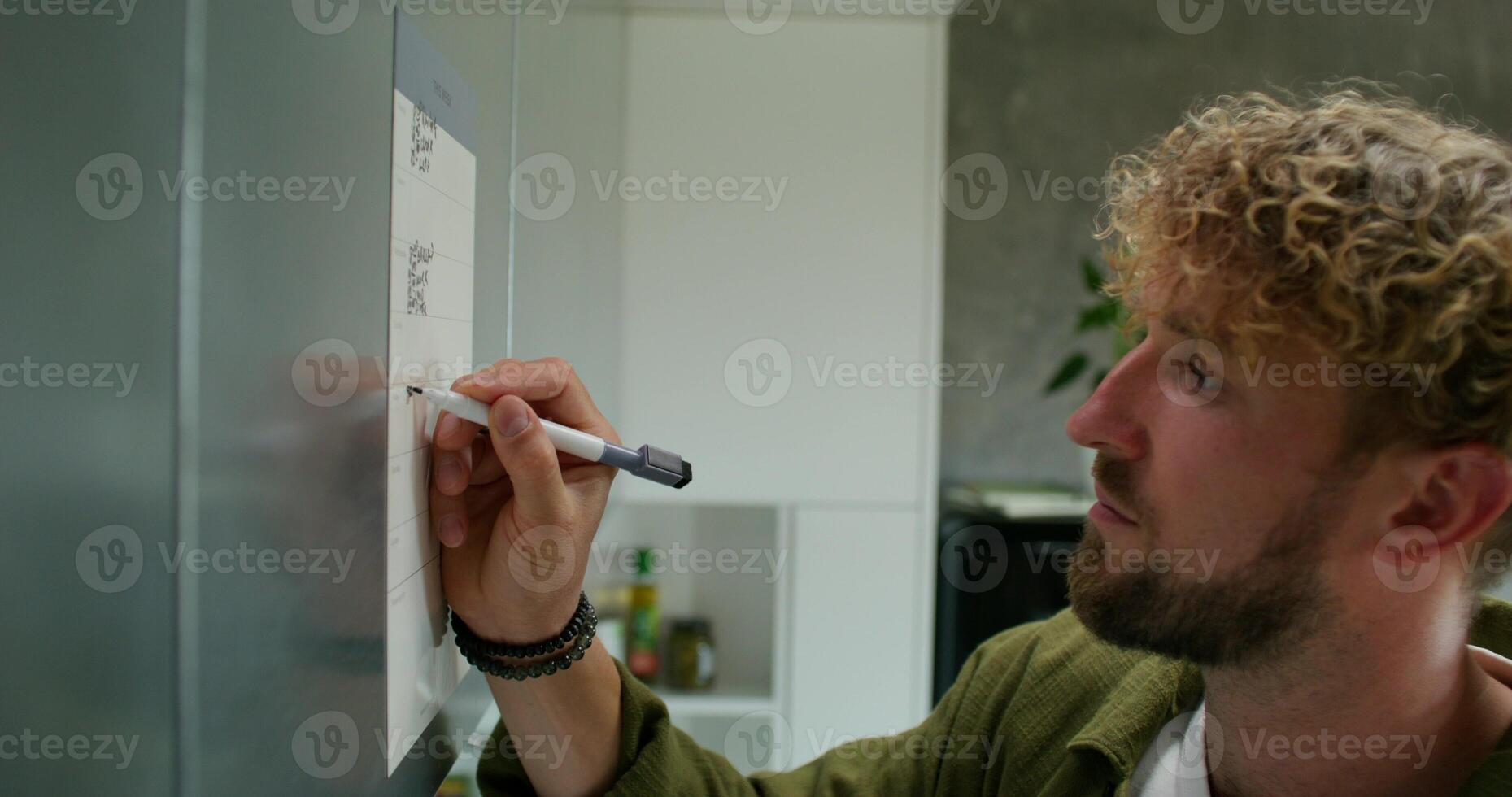 A man in casual clothes fills out a chart with a schedule hanging on the kitchen cabinet. Daylight, home interior - kitchen photo