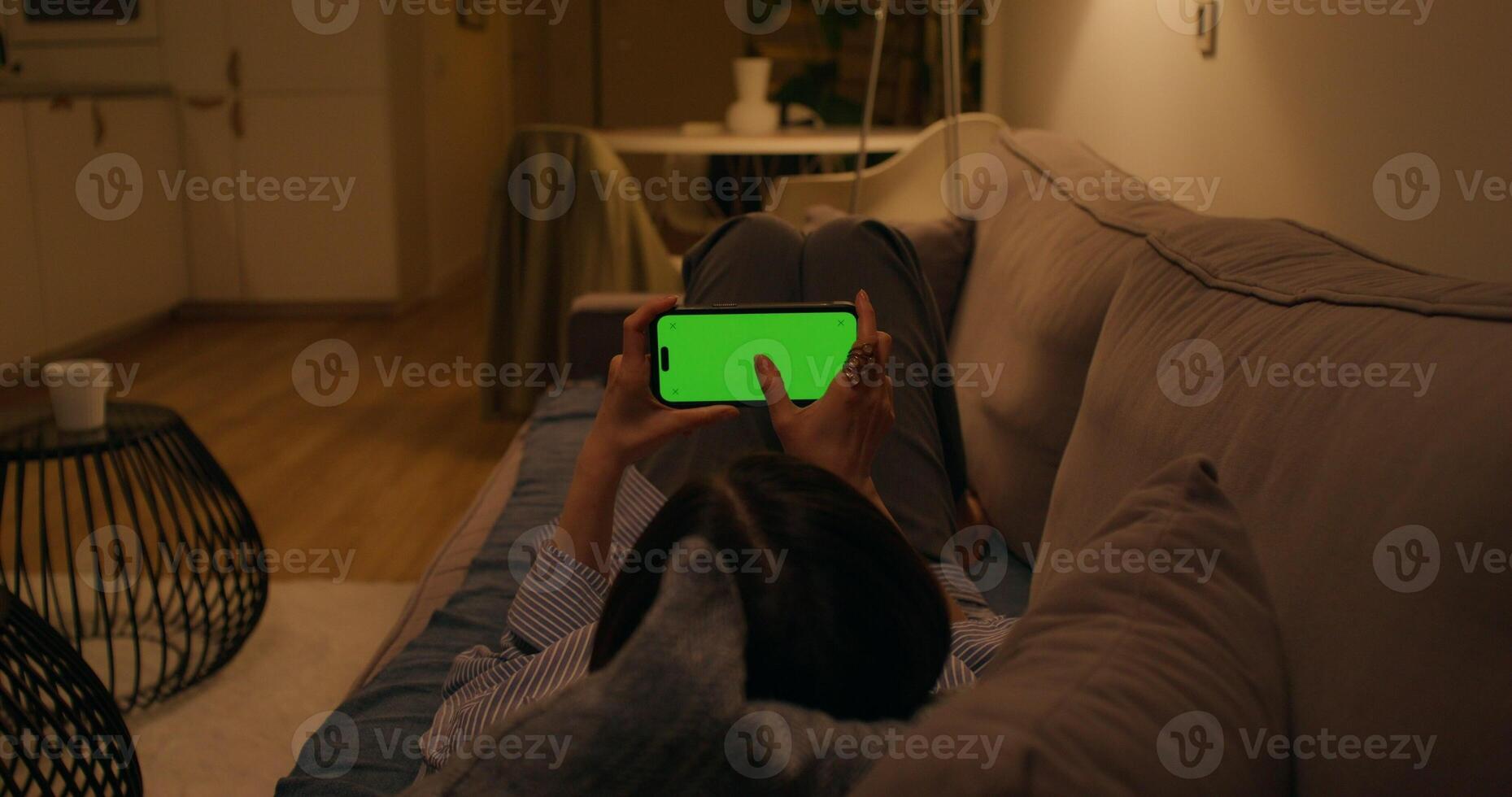 A woman uses a mobile phone with a green screen lying on the couch at home, dimmed evening light - dimmed photo