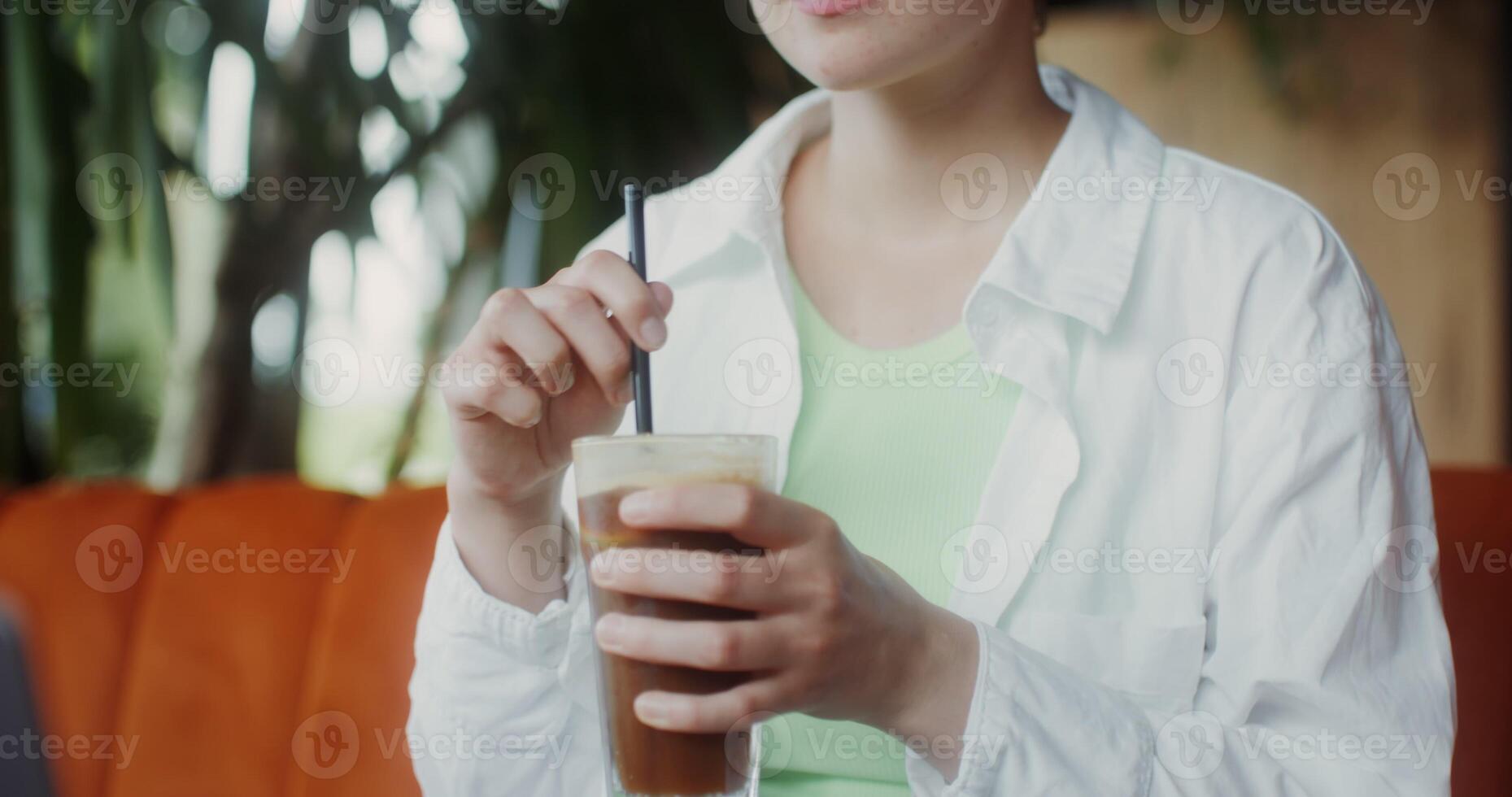A young woman drinks ice coffee from a straw, sitting at a table opposite an open laptop in a cafe, cheesecakes are on the table photo
