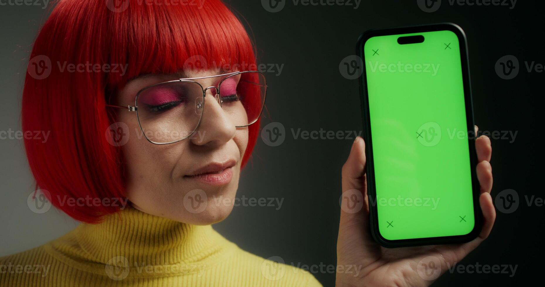 A woman with glasses and an orange wig smiles at the camera, holding a mobile phone with green screen in her hand, standing on a gray background in the studio - holding photo