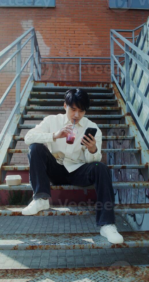 A young man of Chinese appearance uses a mobile phone and, drinks juice from a disposable cup with a straw while sitting on an outdoor metal staircase photo