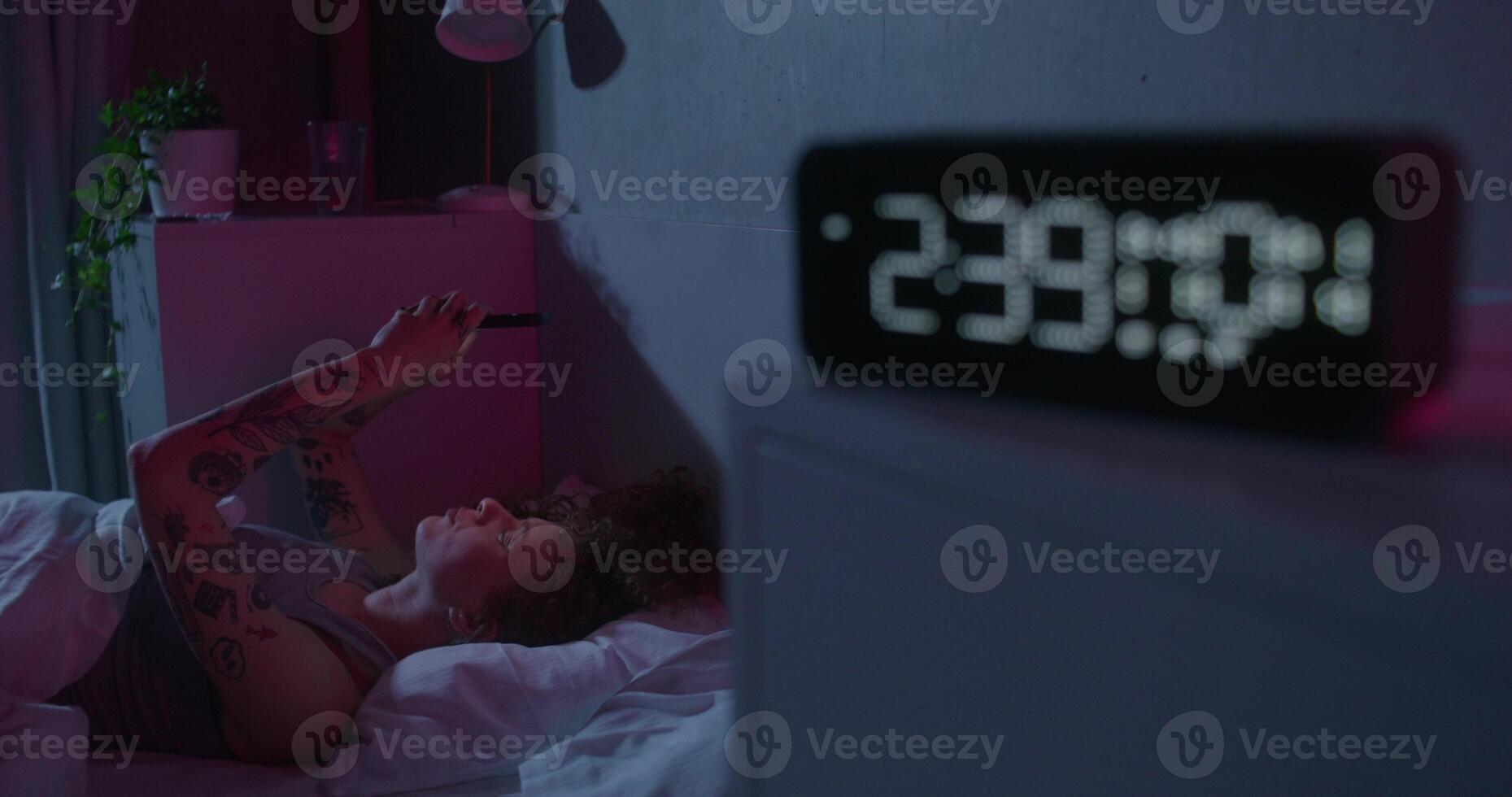 A woman uses a mobile phone lying on the bed. Dark room, home interior. The electronic clock is on the bedside table - bedside photo