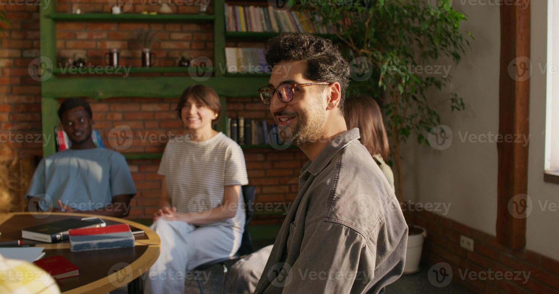 A group of young people are smiling and talking while sitting around a table. They study the Bible together. Daylight photo