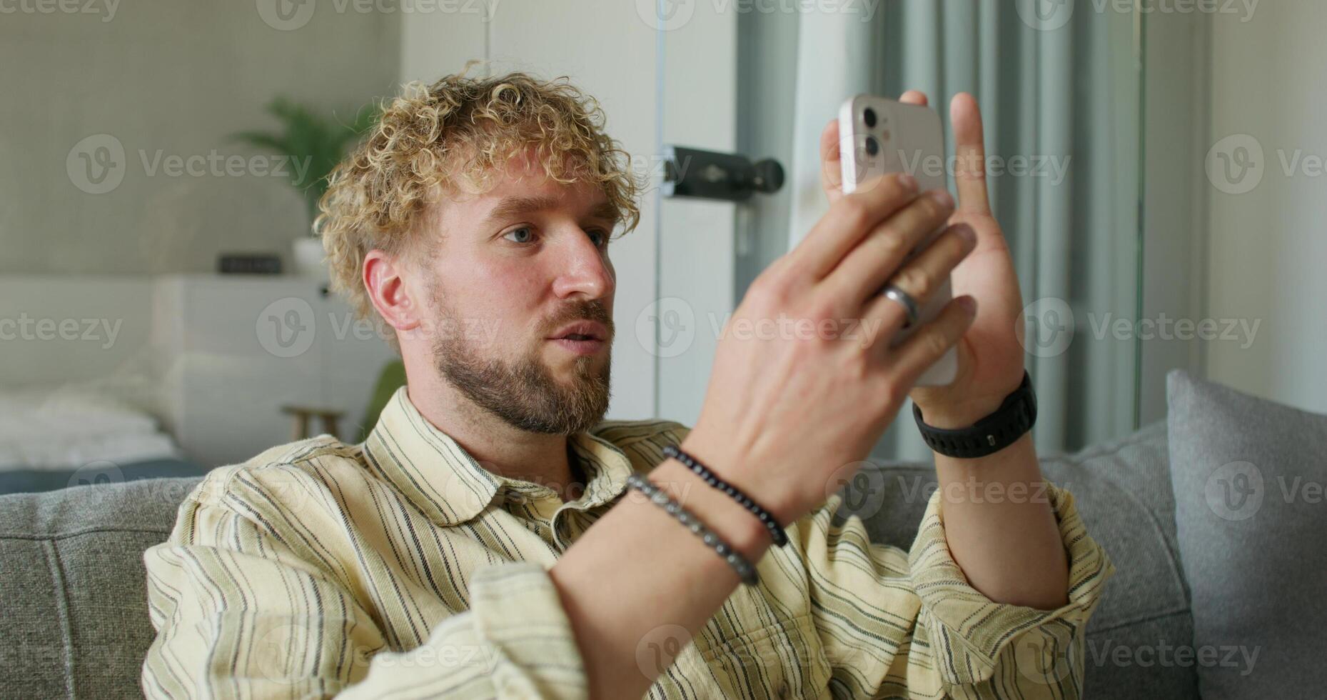 A bearded man in casual clothes is talking on a chat on a mobile phone sitting on the couch at home. Daylight - clothes photo