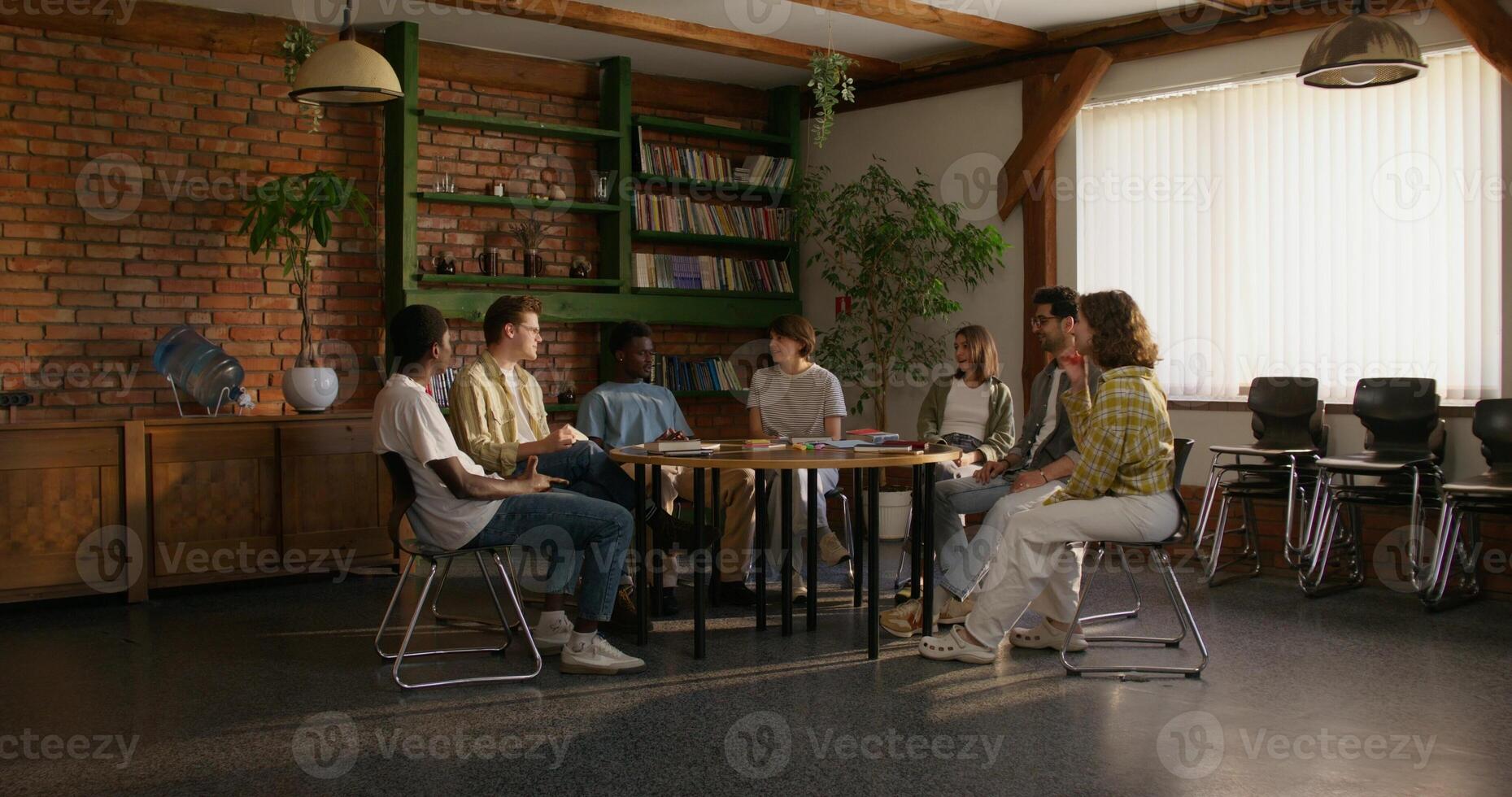 A group of young people are smiling and talking while sitting around a round table indoors. Notebooks and books on the table. Daylight - sitting photo
