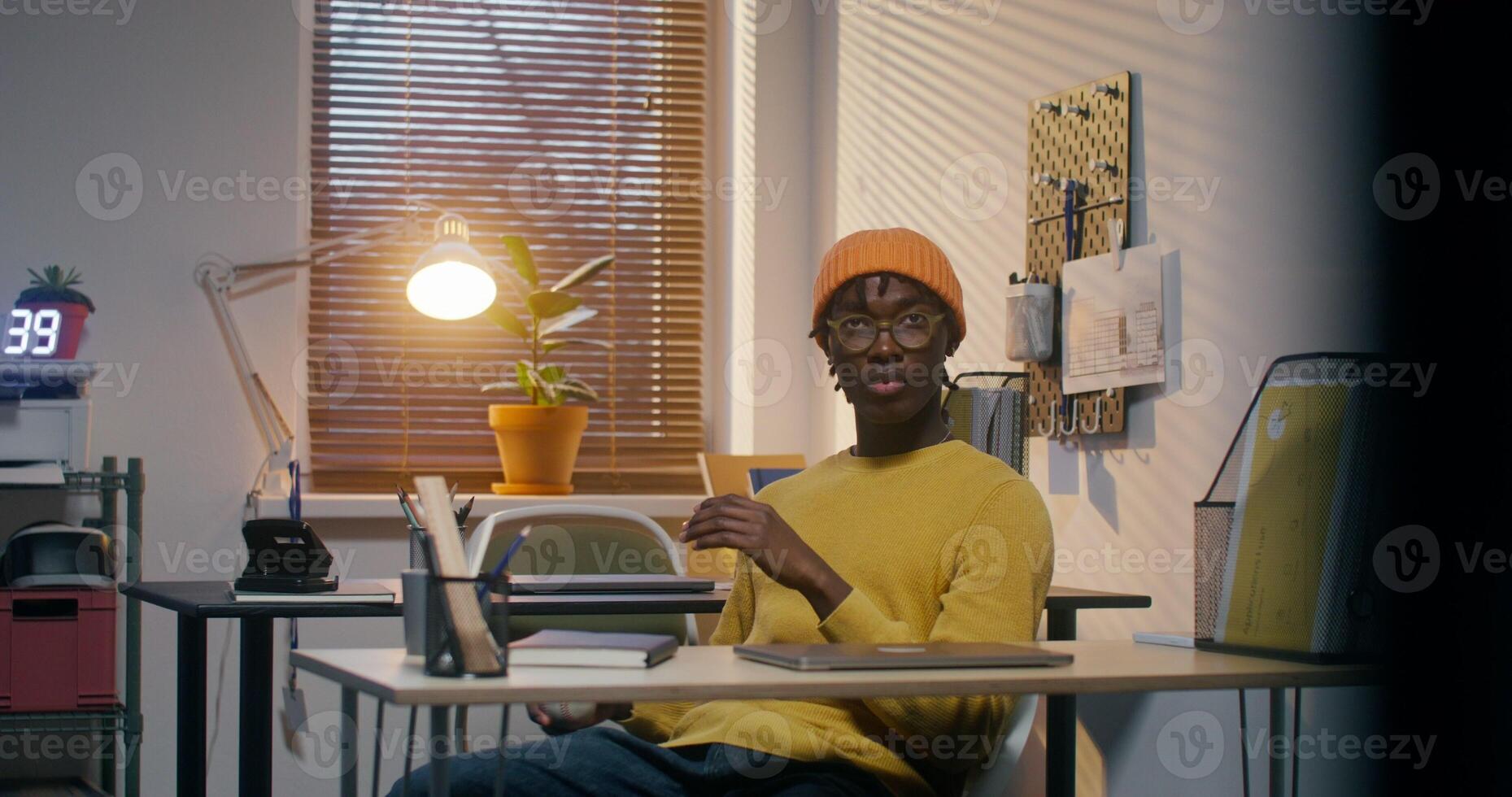A man with glasses and a knitted hat is playing with a baseball and typing on a laptop sitting at a desk in the office - playing photo