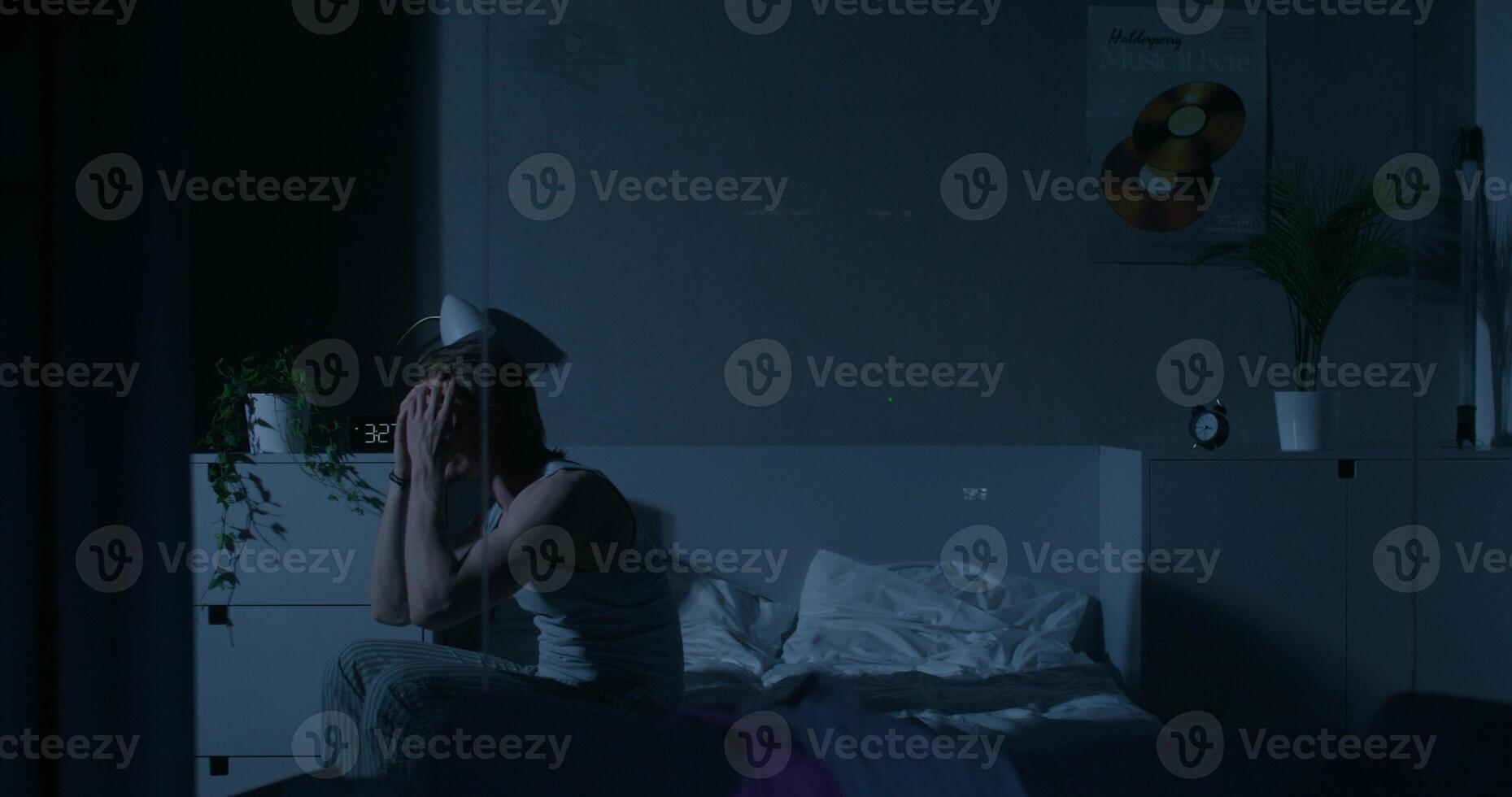 A man rubs his face while sitting on the bed, he can't sleep. Home interior, dark room photo