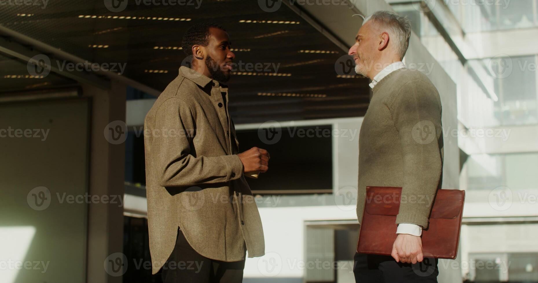A white-haired European man with a laptop in a case and a young African American man with a disposable cup communicate with each other in the city center. in 4k, red komodo photo