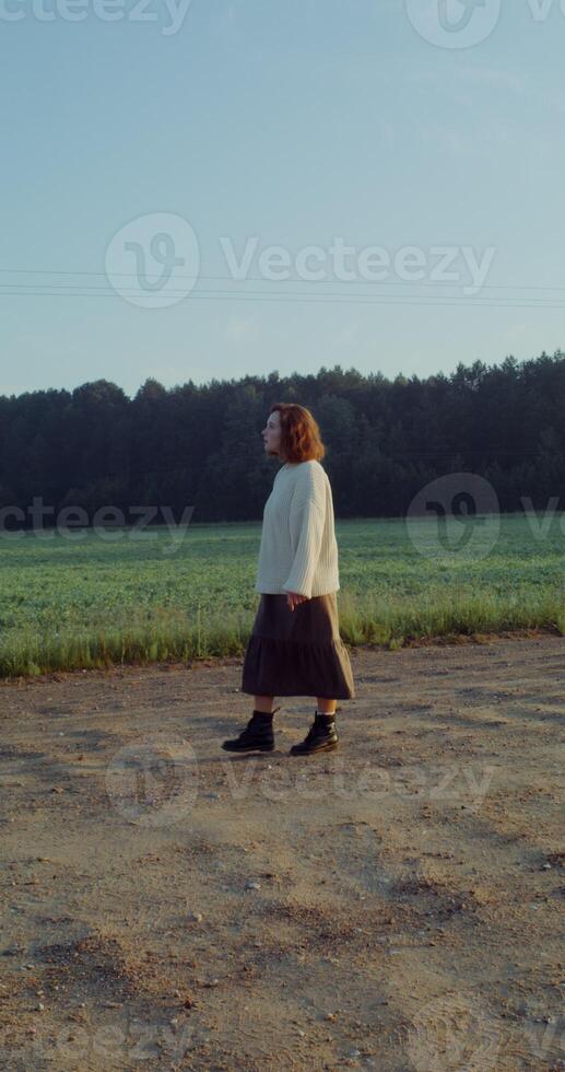 A young, red-haired, warmly dressed woman surveys her surroundings as she walks along a gravel agricultural road in the rays of the rising sun. 4k, vertical , red komodo - surveys photo