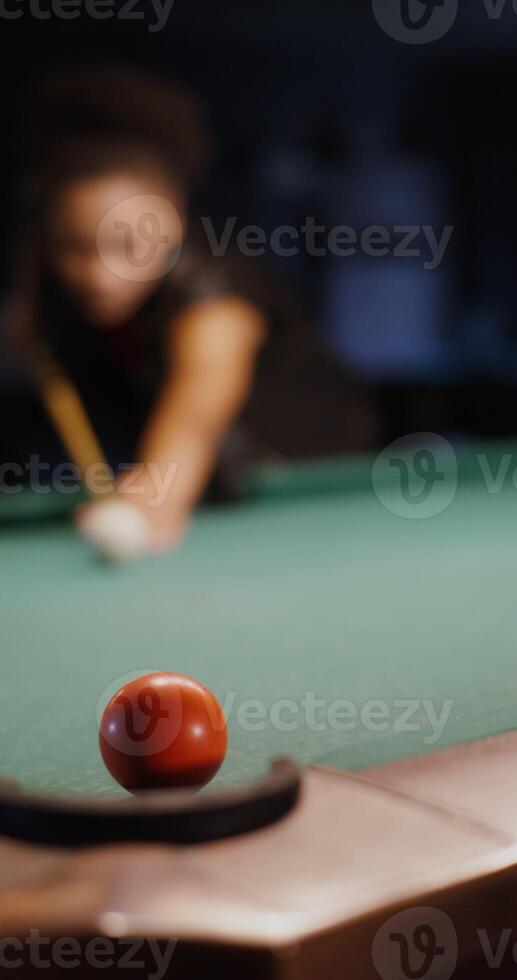A young beautiful African-American girl is intently aiming a cue at the ball while playing billiards, but she fails to hit the hole, vertical photo