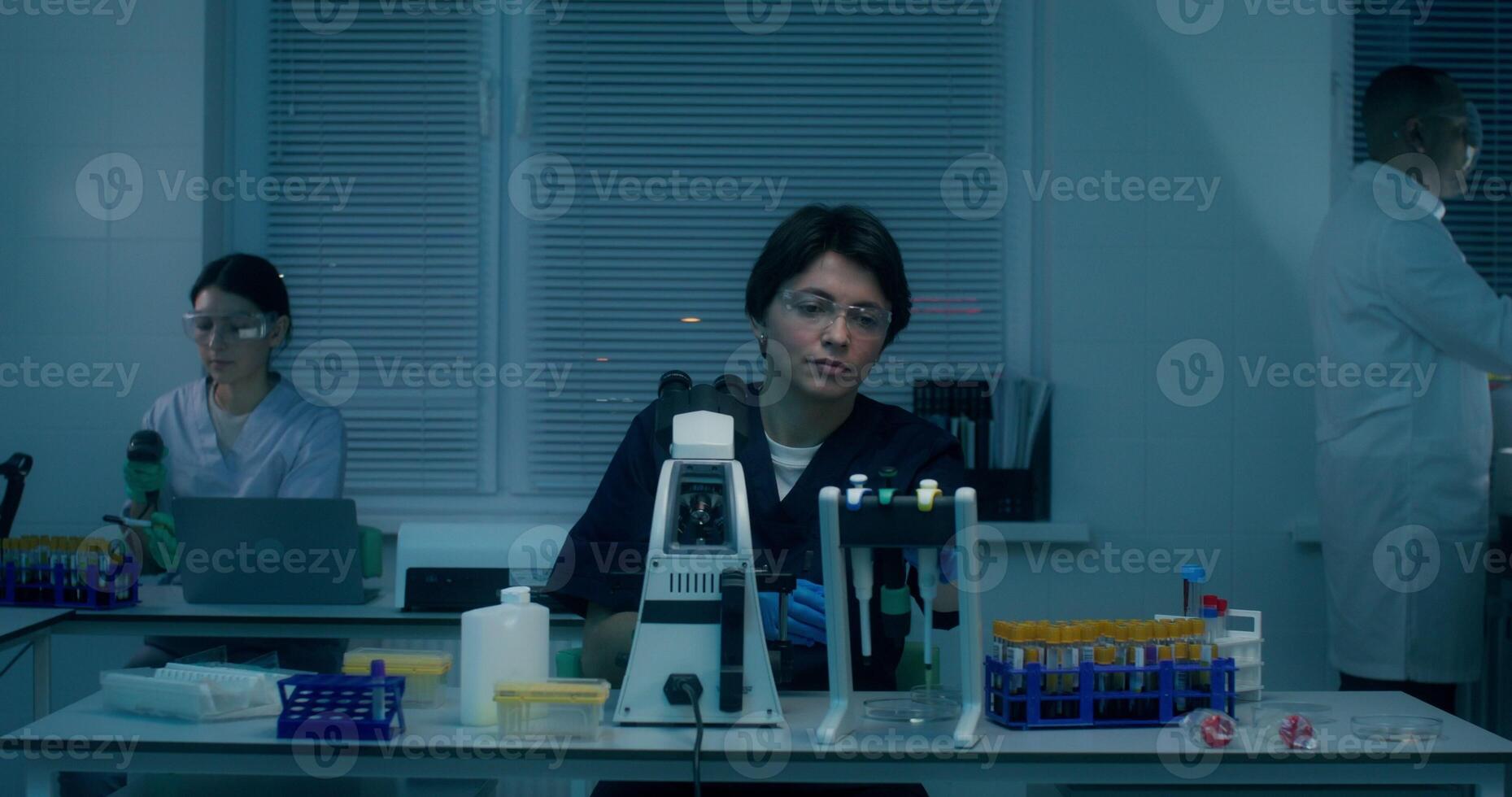 A group of laboratory assistants in medical clothes work in a medical laboratory, using a microscope and conducting a blood test - medical - of photo