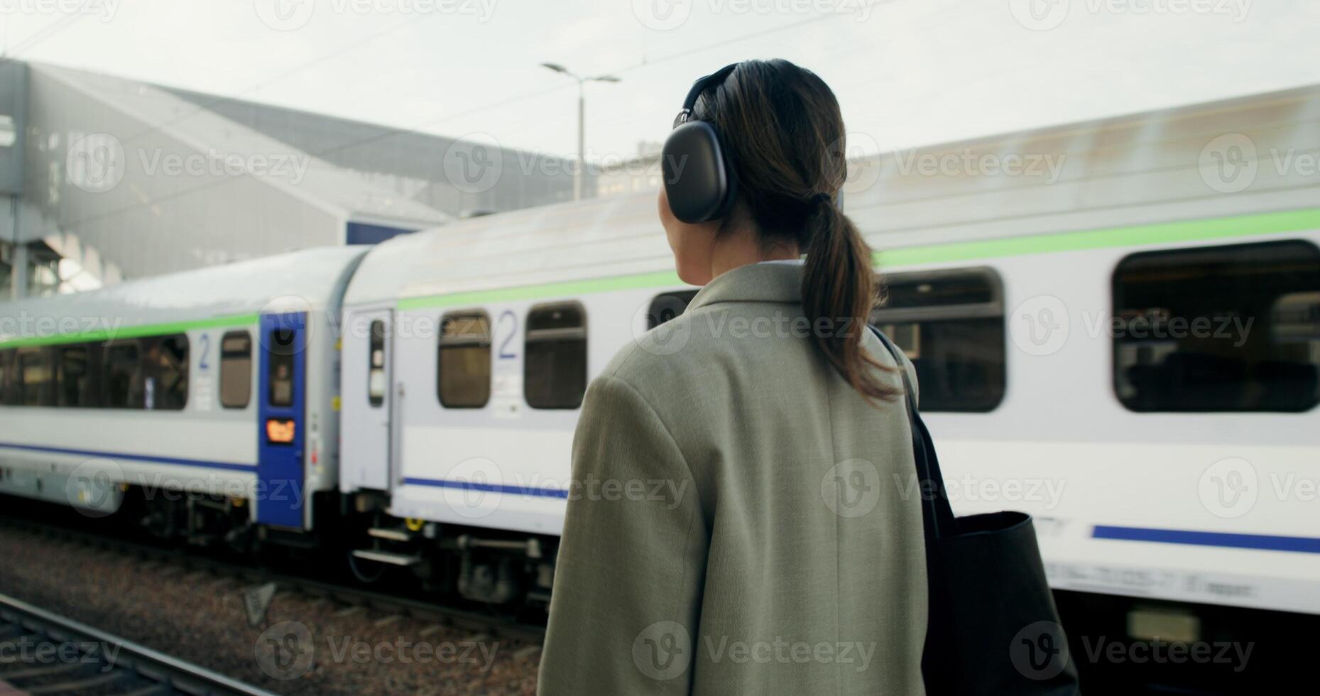 A woman in formal clothes and with headphones on her head uses a mobile phone and drinks coffee from a disposable cup walking along the railway platform - coffee photo