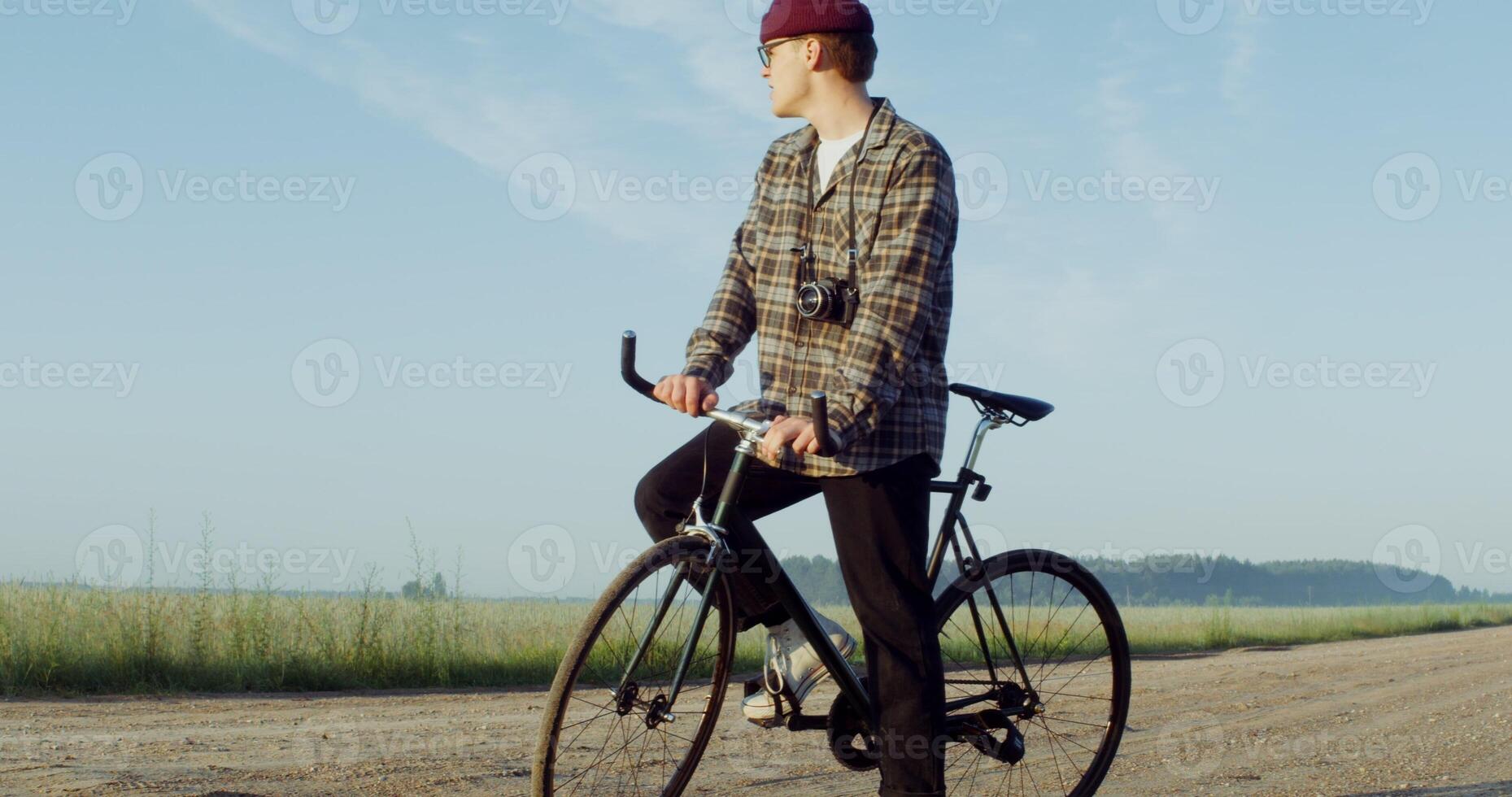 A young stylishly dressed man with a camera hanging around his neck stands in the middle of a gravel road with a bicycle in his hands and admires the fields around him. 4k , red komodo photo