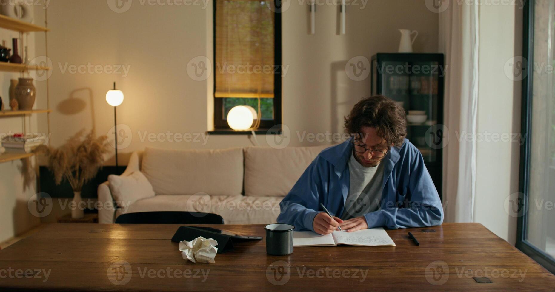A man with glasses is writing in a notebook sitting at a table with a serious face. Stylish home interior - notebook photo