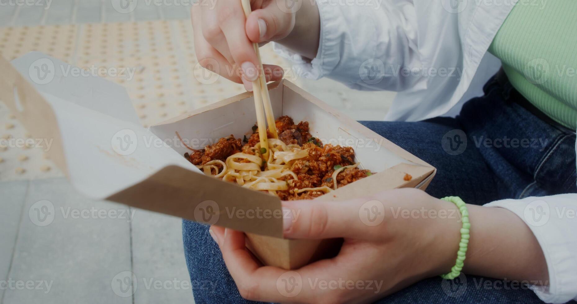 A woman eats noodles out of a box using chopsticks sitting outdoors, close photo