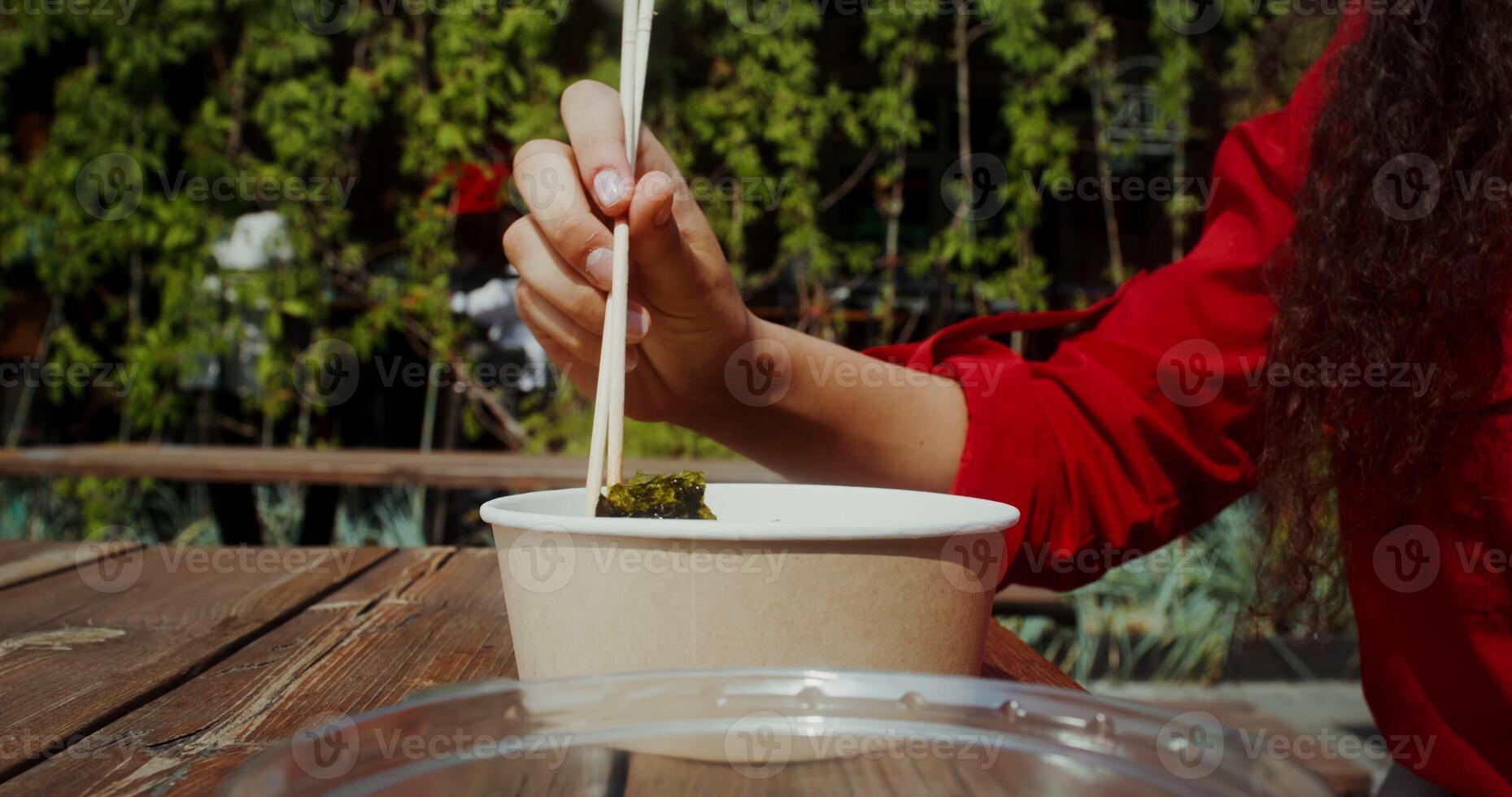 A young woman eats Asian food out of a box using chopsticks while sitting at a table on the cafe terrace, hands close-up, no face - chopsticks photo