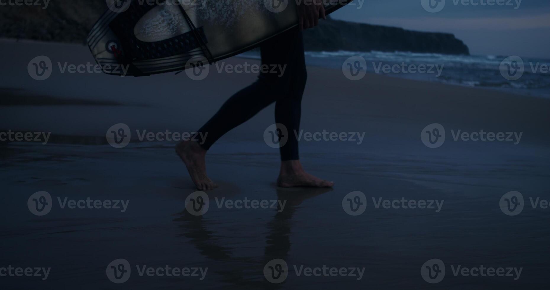 A bearded man with a surfboard in his hands walks along the seashore at dusk photo