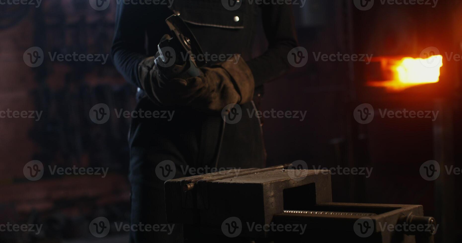 A young blacksmith of African American appearance in goggles and an apron turns on an angle grinder and begins to cut off a piece of metal workpiece while working in a forge. 4k, red komodo photo