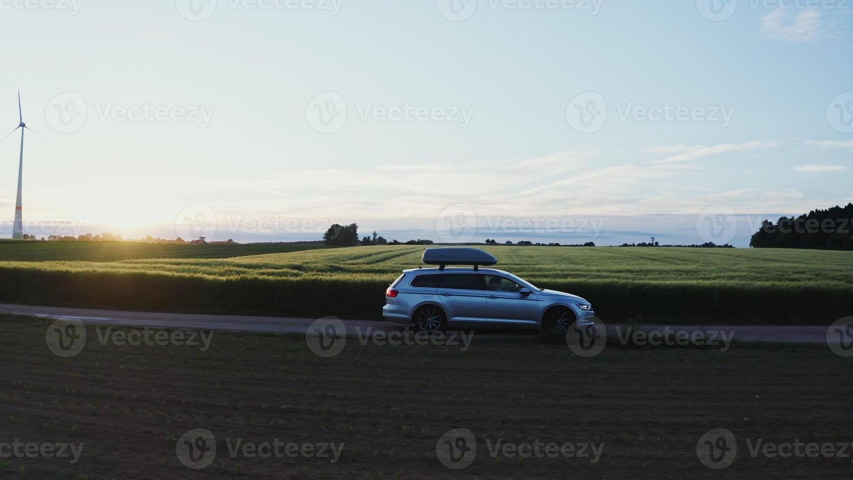 A car with a roof rack drives along a road through picturesque green meadows and arable fields on a beautiful sunny morning. Wind generator in the rays of the rising sun - picturesque photo