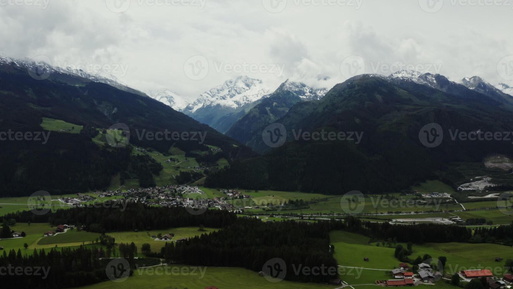 Panoramic view of a picturesque mountain valley with a village in a lowland and rocky mountains, the tops of which are covered with snow, located behind green hills. Alps, Austria. - mountain photo