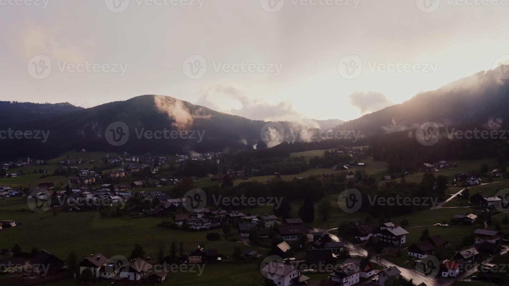 Panoramic view of the cottage town, located in a lowland between the Alpine mountains, the tops of which are covered with white clouds. Alps, Austria, drone - located photo