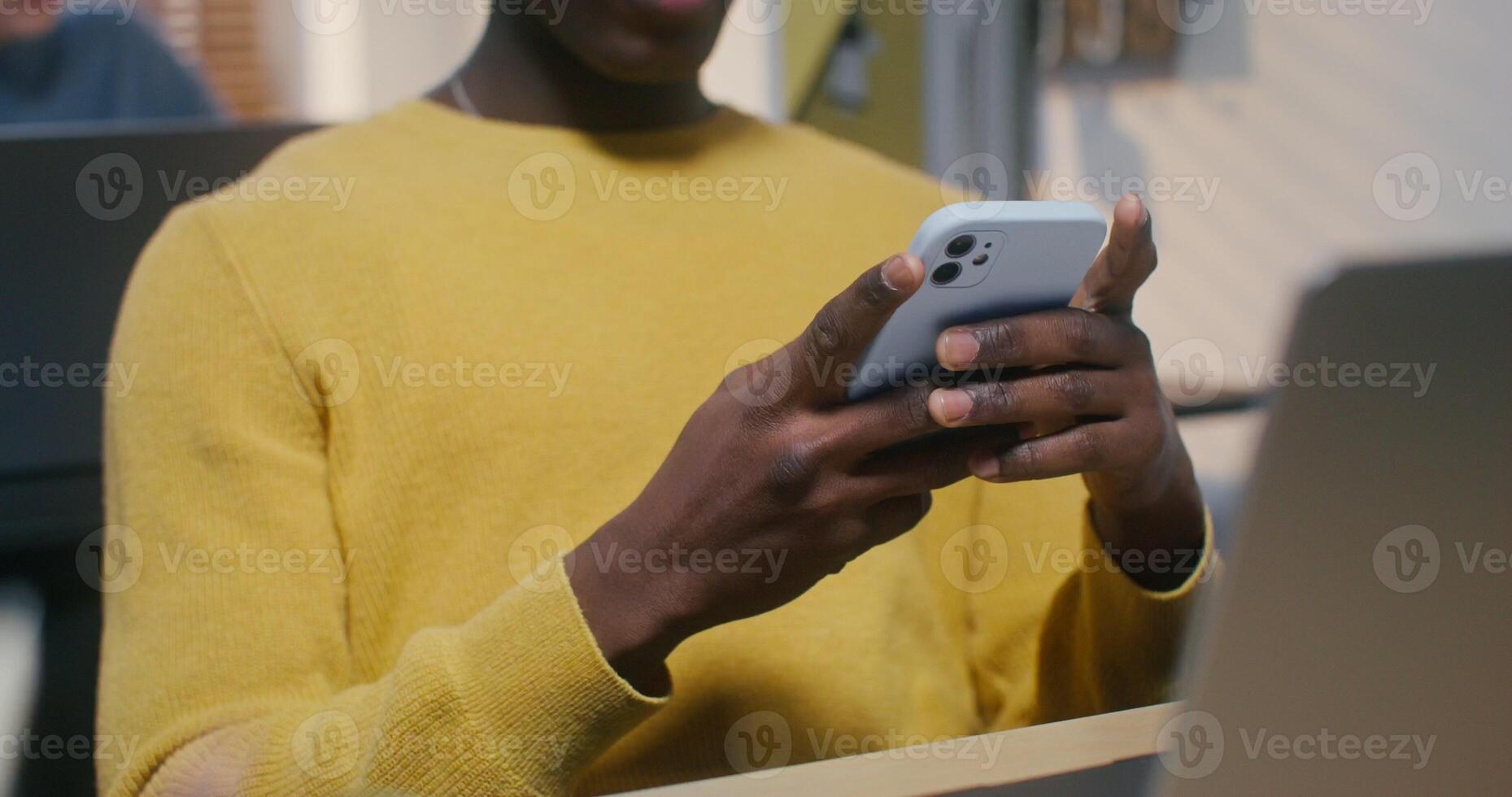 A man in a hat and headphones uses a mobile phone sitting at a desk in the office. His colleague is typing on a laptop sitting behind photo