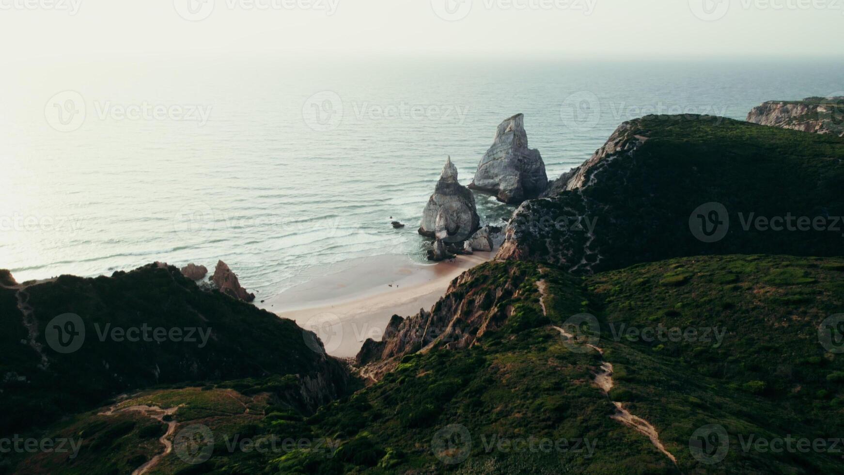 The drone flies from the top of a mountain overgrown with grass towards the sea, with a sandy beach between the rocks - towards photo