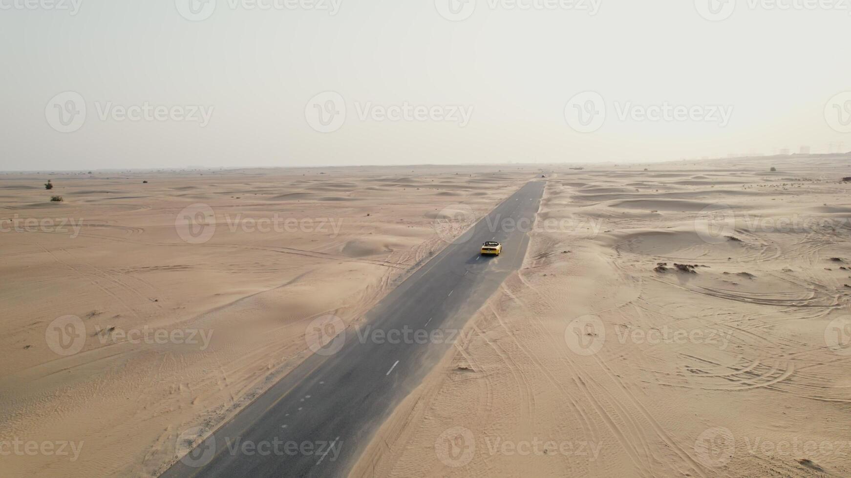 A yellow car is driving along an asphalt road in the desert, raising sand dust behind it. Drone - asphalt photo