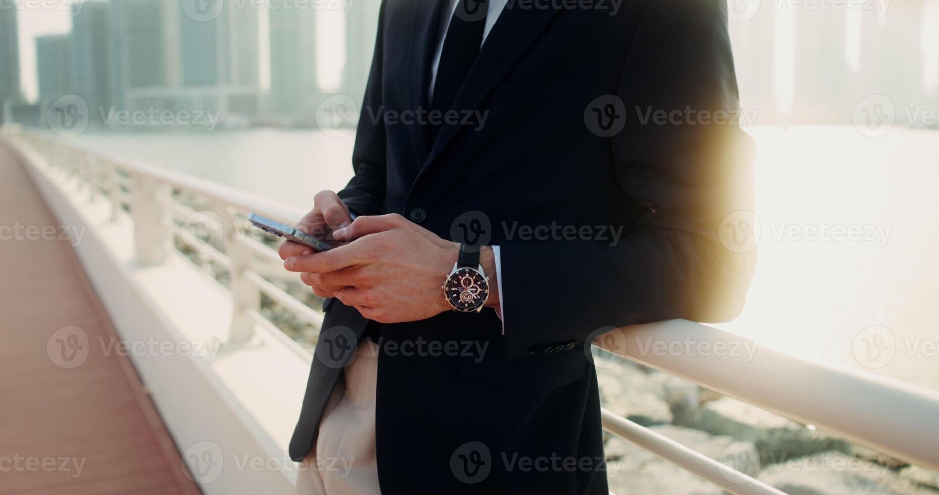 A man in a business suit with a wristwatch on his hand uses a smartphone standing on the embankment on a sunny day. Men's hands close-up - smartphone photo